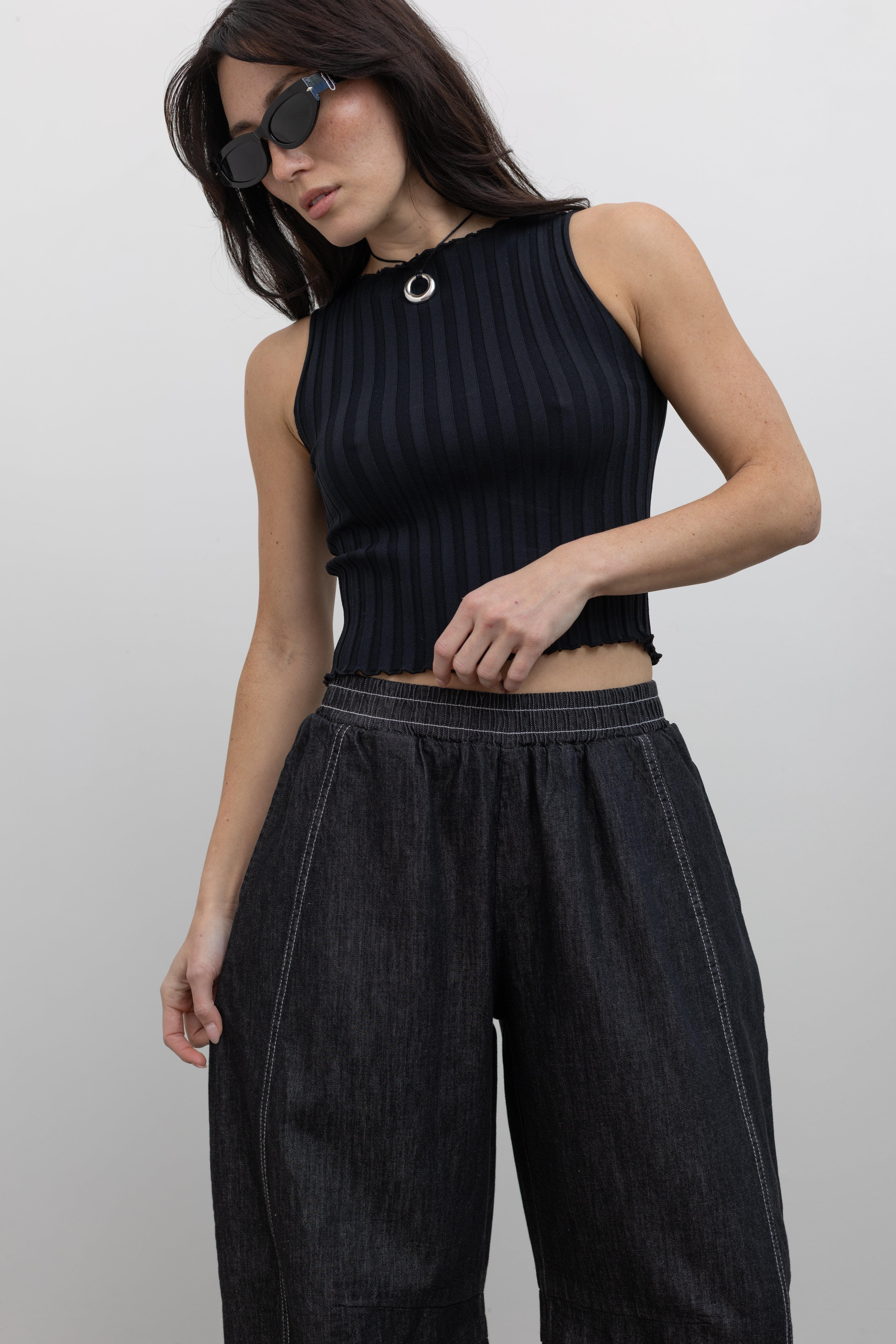 Close-up front view of a woman standing wearing elevated and chic Alas Eius label black cotton barrel leg pants featuring contrasting white double stitching, side seam pockets, and an elastic waistband paired with a black ribbed knit crop tank and ATP black leather cut out sandals at an Alas Eius store. 