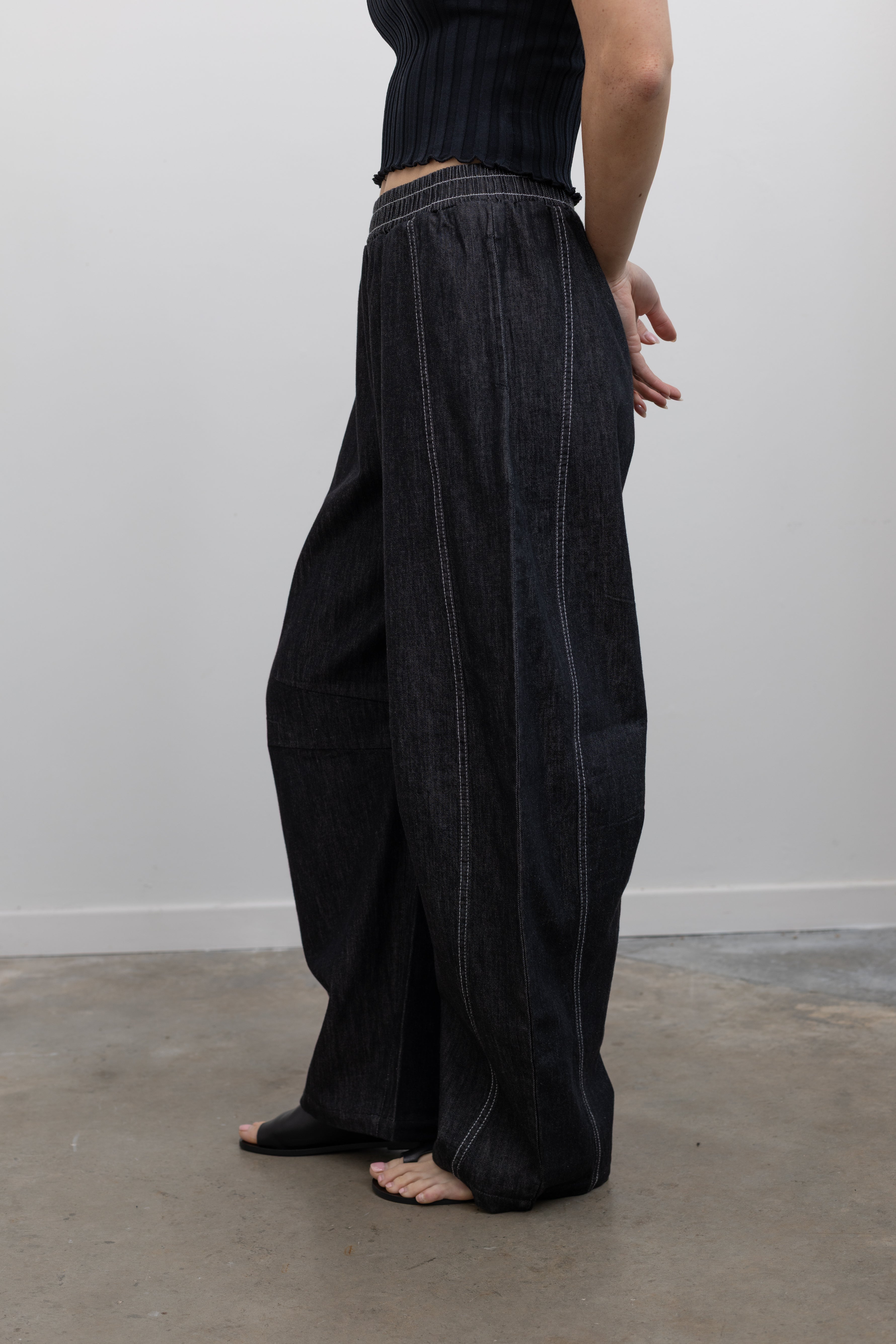 Close-up side view of a woman standing wearing elevated and chic Alas Eius label black cotton barrel leg pants featuring contrasting white double stitching, side seam pockets, and an elastic waistband paired with a black ribbed knit crop tank and ATP black leather cut out sandals at an Alas Eius store. 