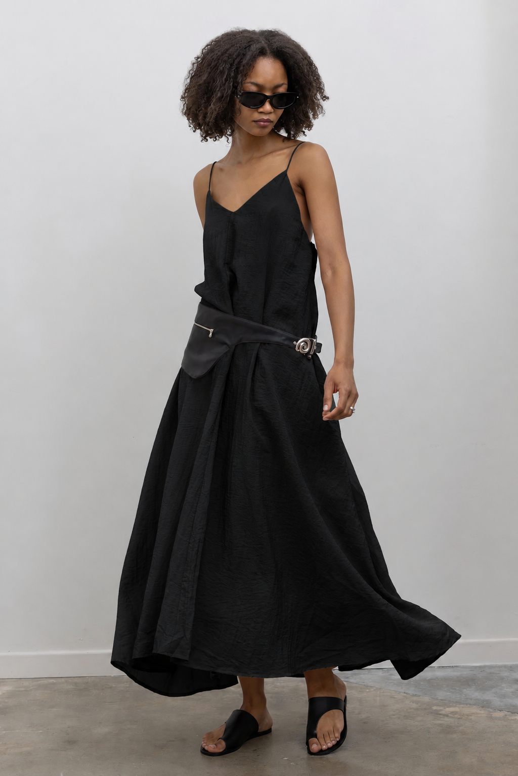 Full length front view of a woman wearing an elegant yet minimalist Alas Eius label spring and summer dress in a black cotton blend featuring a flowing silhouette, adjustable bungee straps for a customized fit and two convenient side pockets paired with Nomia black leather zip belt bag around her waist and black leather sandals at an Alas Eius store. 