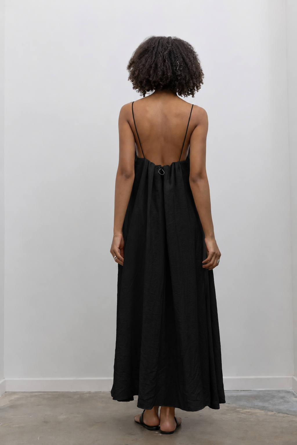 Full length back view of a woman wearing an elegant yet minimalist Alas Eius label spring and summer dress in a beige cotton blend featuring a flowing silhouette, adjustable bungee straps for a customized fit and two convenient side pockets paired with Nomia black leather zip belt bag around her waist and black leather sandals at an Alas Eius store. 