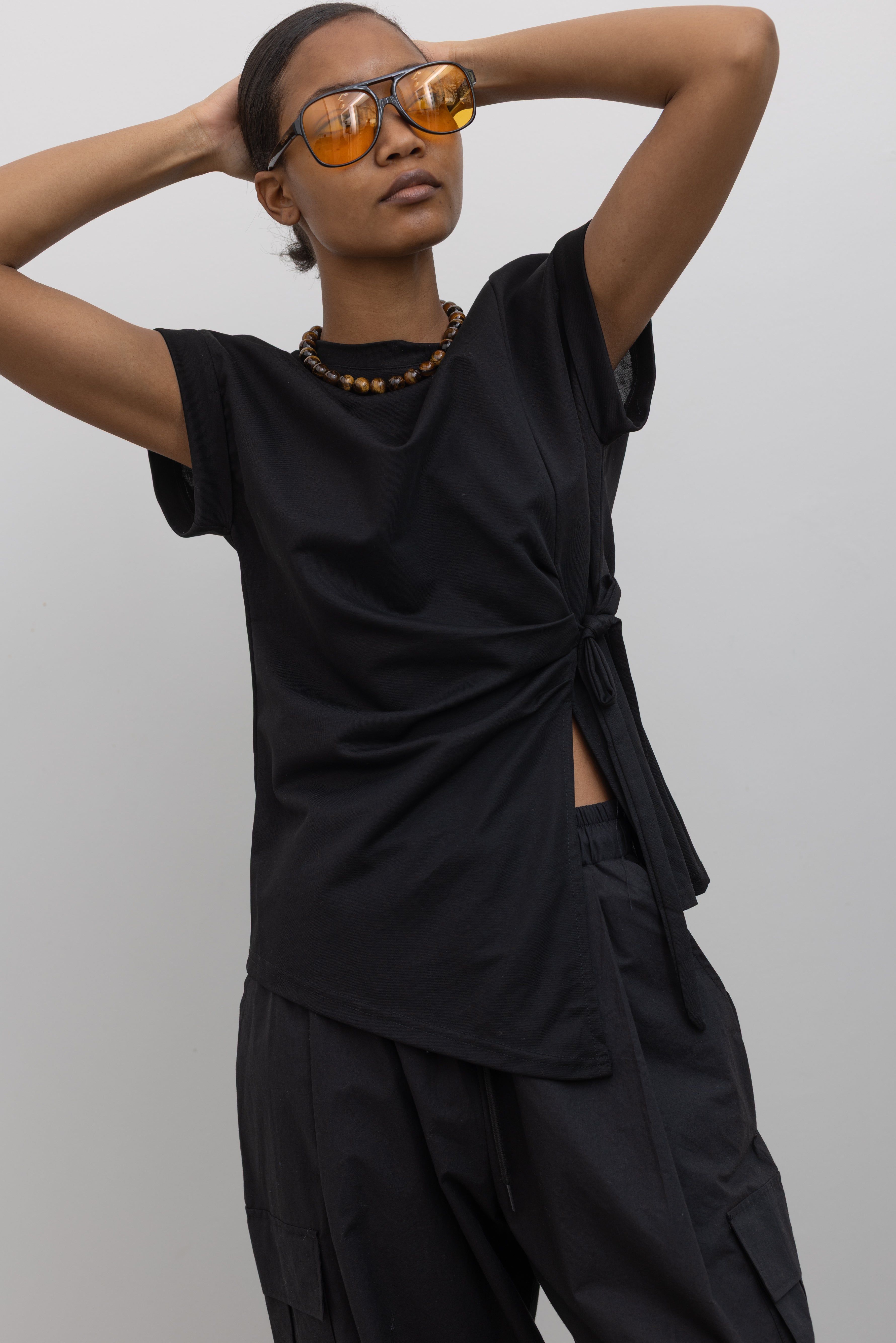 Woman wearing essential capsule wardrobe staple in an Alas Eius Black Drop Shoulder Ribbon Side Tie Cotton Top with black cargo pants and sunglasses at Alas Eius