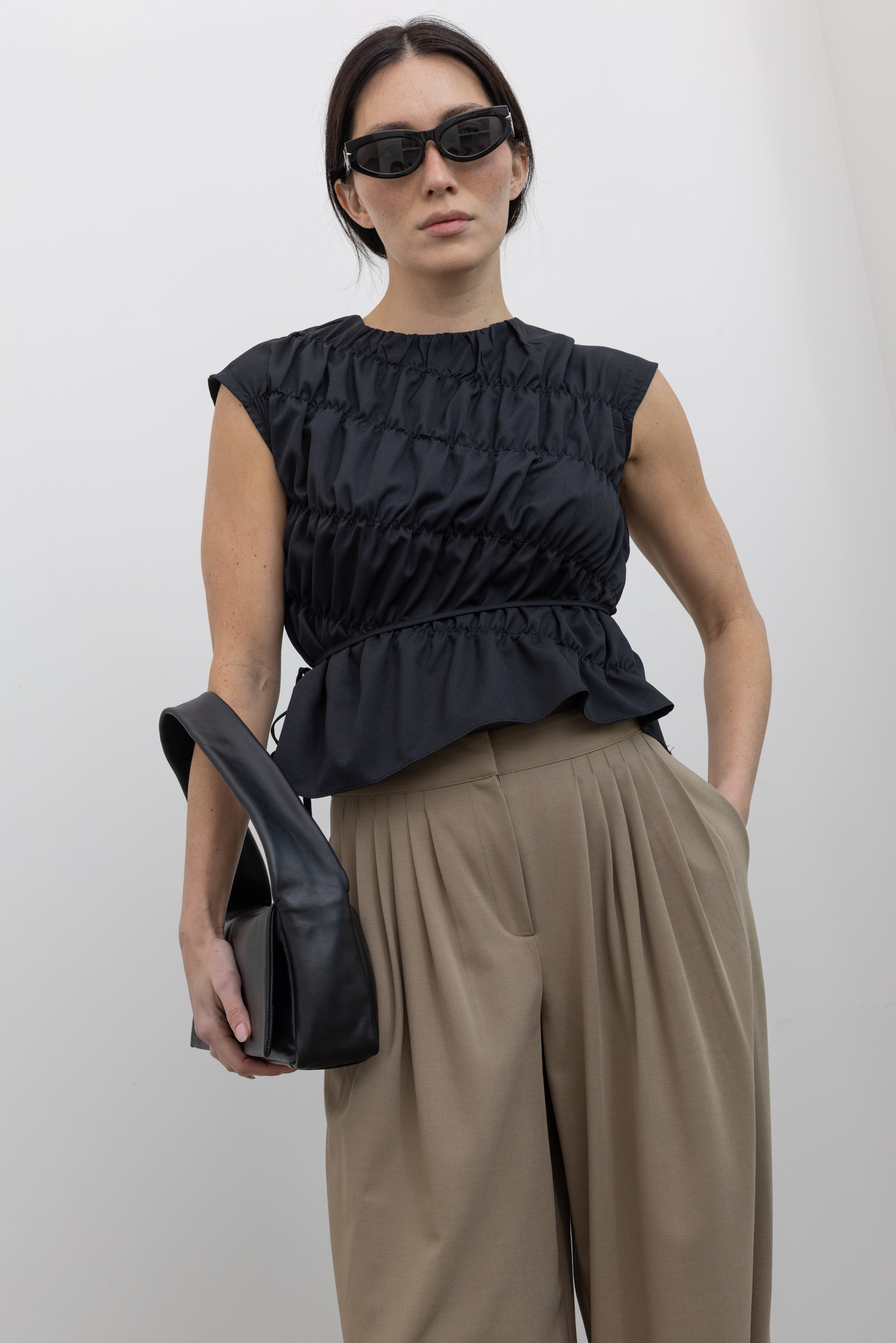 Front view of a woman posing wearing contemporary and chic women's fashion in an Alas Eius Black Asymmetrical Cinched Top featuring a crew neckline, cap sleeves, and an optional waist tie paired with elevated khaki cinched ankle pants, silver sandals, and a black leather St. Agni bag at an Alas Eius store. 