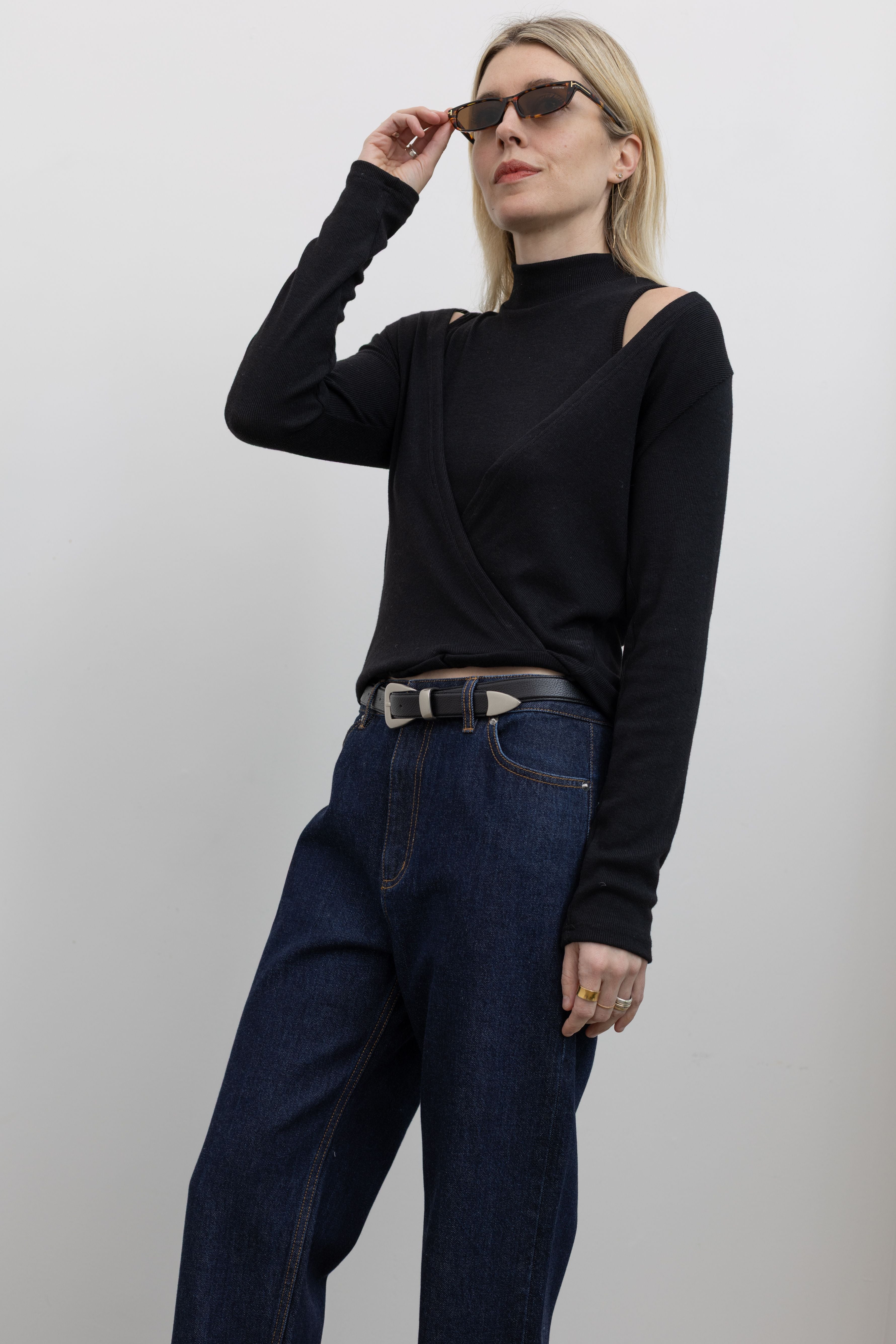 Side view of Woman wearing Alas Euis label deep v-neck double layer long sleeve black knit top with mock neck and open shoulders with dark wash Tibi jeans at alas eius