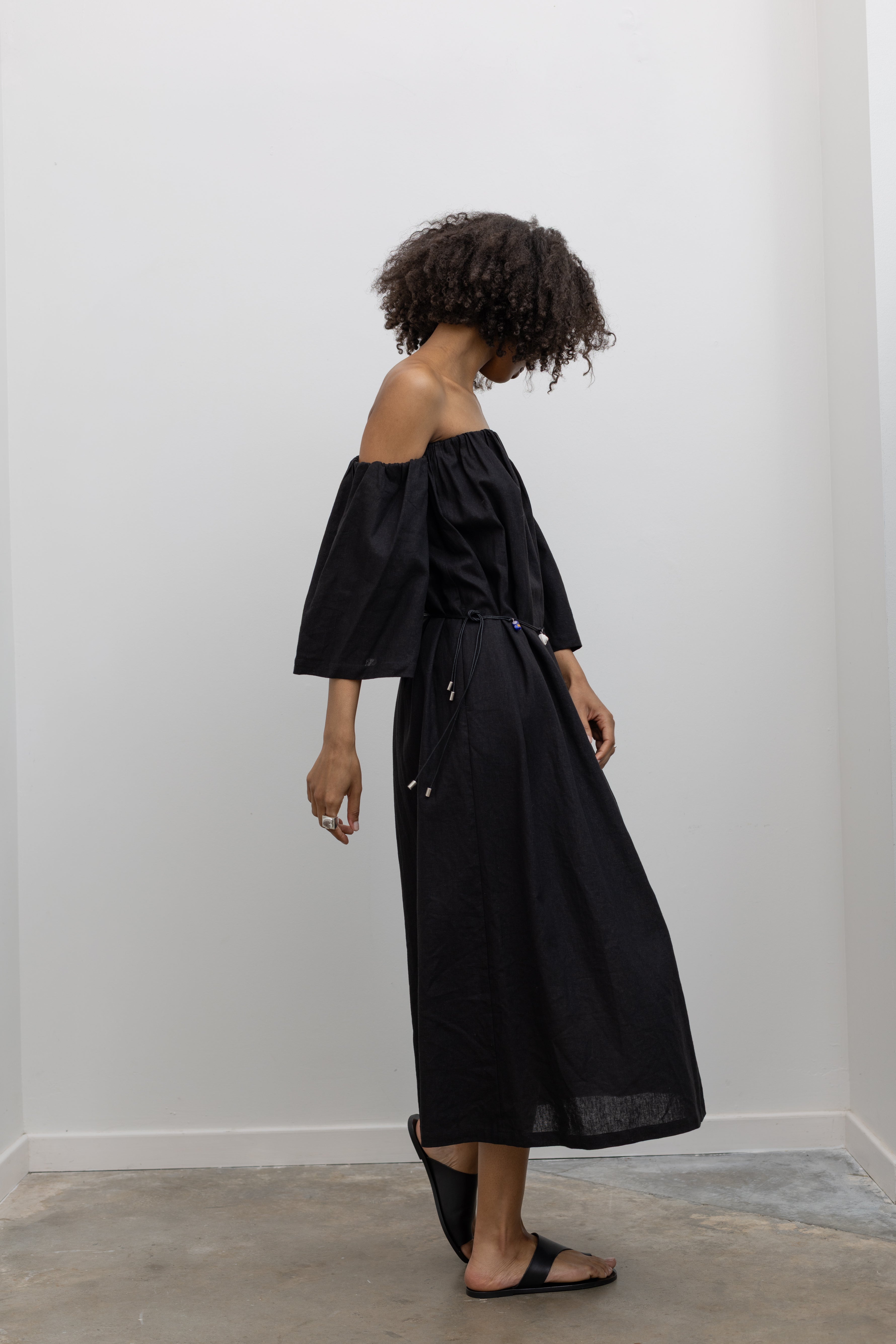 Side view of a woman in motion wearing chic summer fashion in a Alas Eius label black linen midi dress featuring removable sleeves paired with a beaded belt at an Alas Eius store.