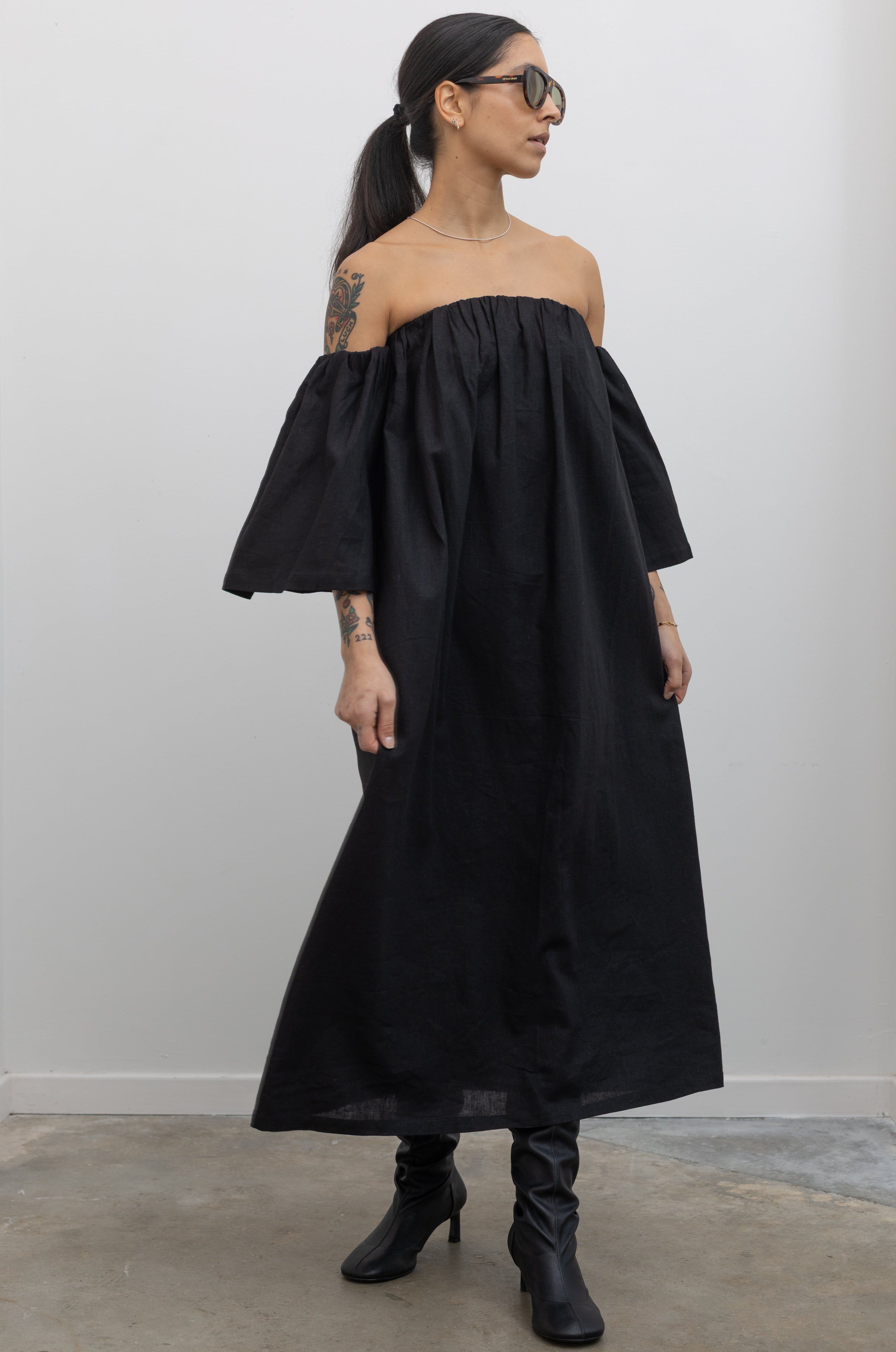 Woman standing wearing fashion in a Alas Eius Label Black Linen Off The Shoulder Midi Dress with black leather boots at Alas Eius
