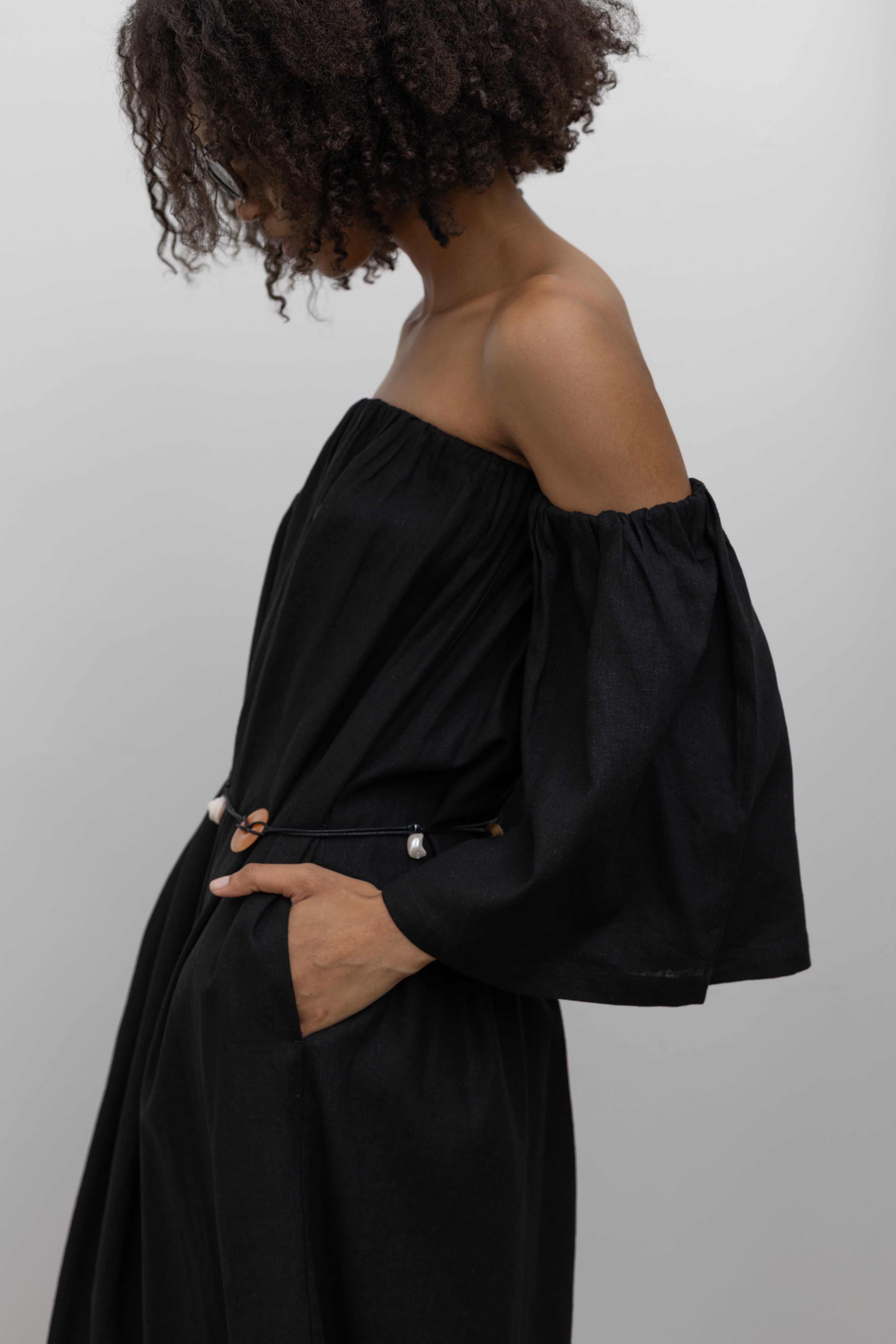 Side view of a woman standing with her hands in the pockets of a chic Alas Eius label black linen midi to maxi dress featuring removable bell sleeves paired with a beaded belt at an Alas Eius store.