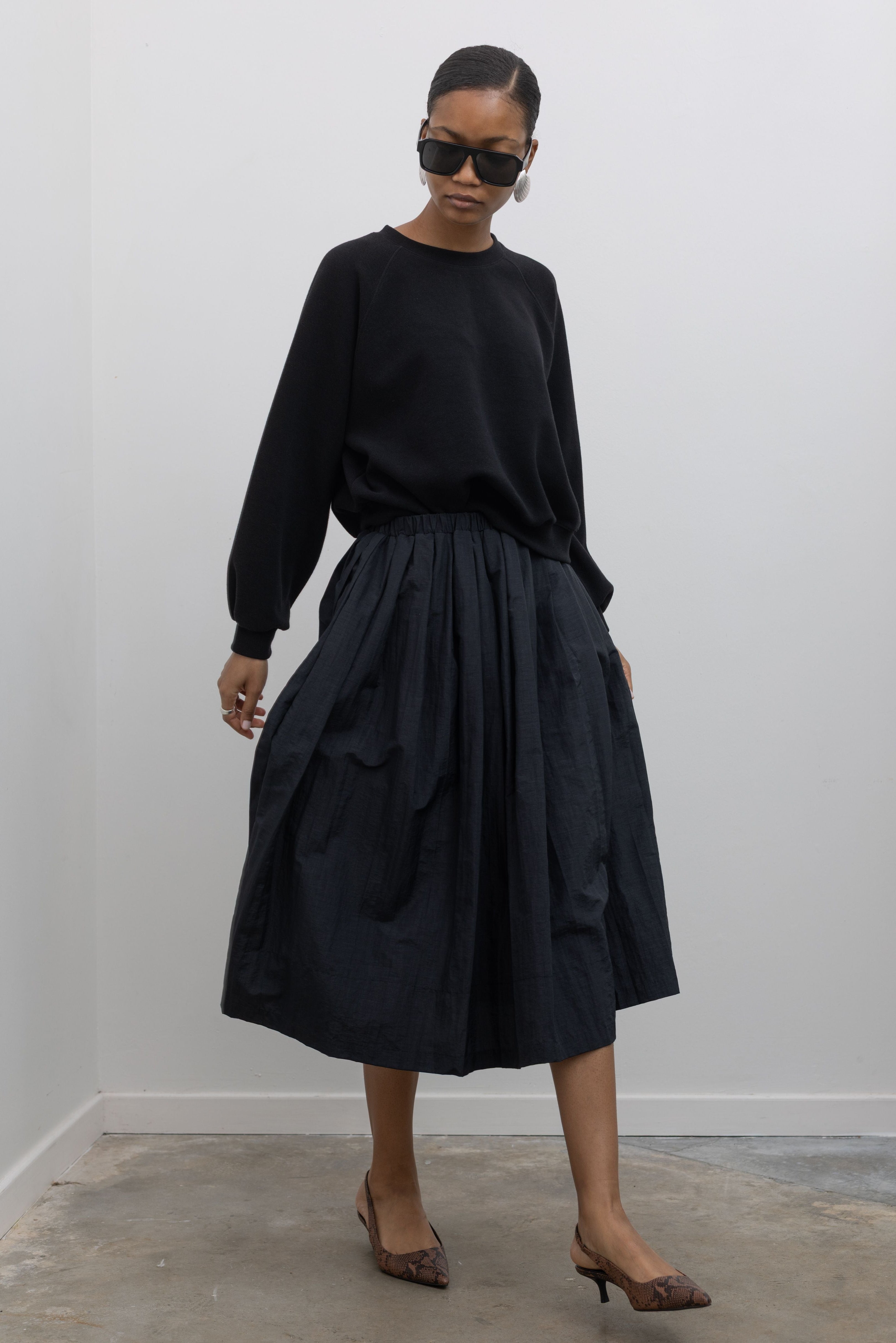 Woman in motion wearing a refined styled outfit including a sophisticated Alas Eius Label black lightweight nylon pleated midi skirt, featuring a fully lined silhouette and finished with an elastic waistband and convenient side pockets. 