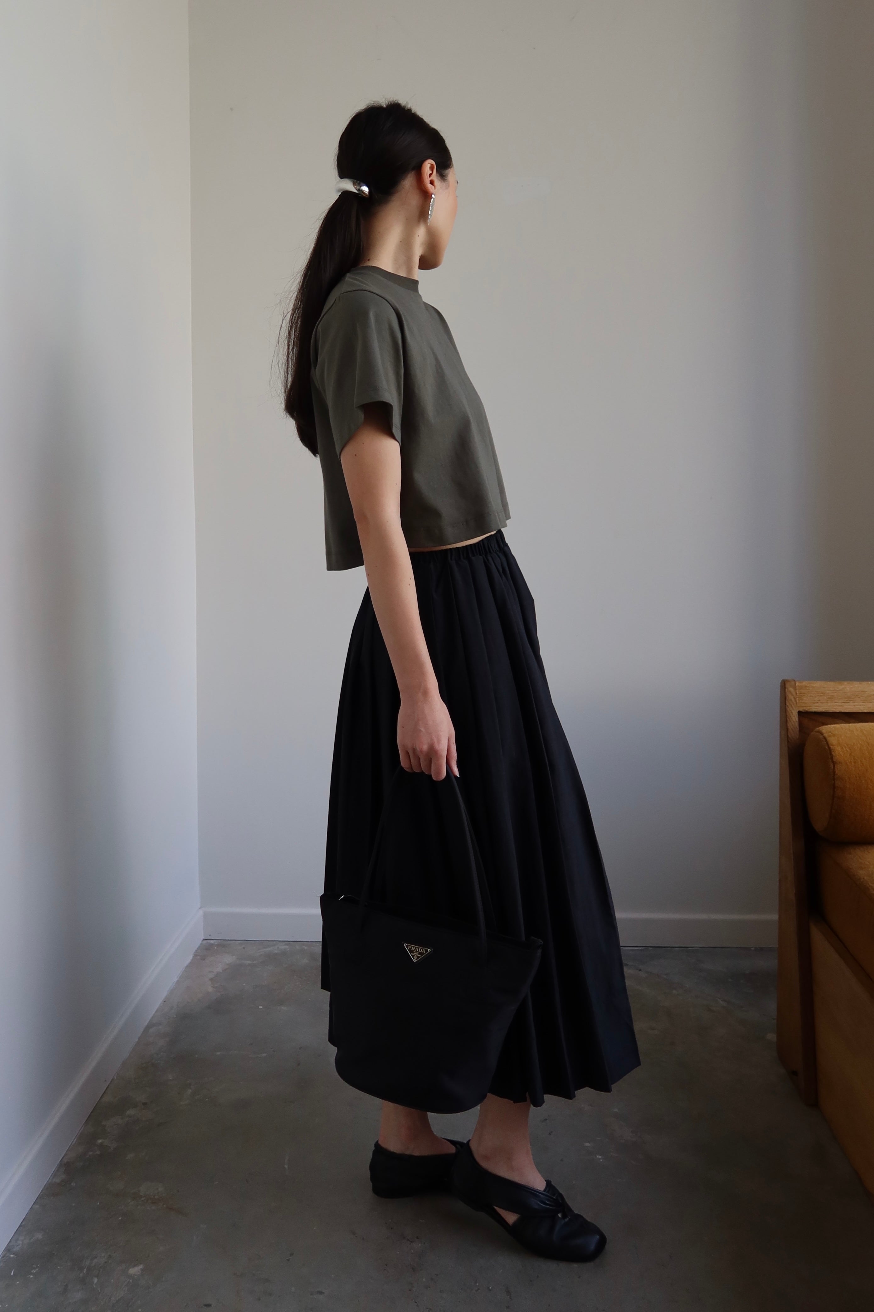 Side view of a woman standing looking over her shoulder wearing an Alas Eius black nylon pleated midi skirt paired with black leather flats and a cropped t-shirt at Alas Eius. 