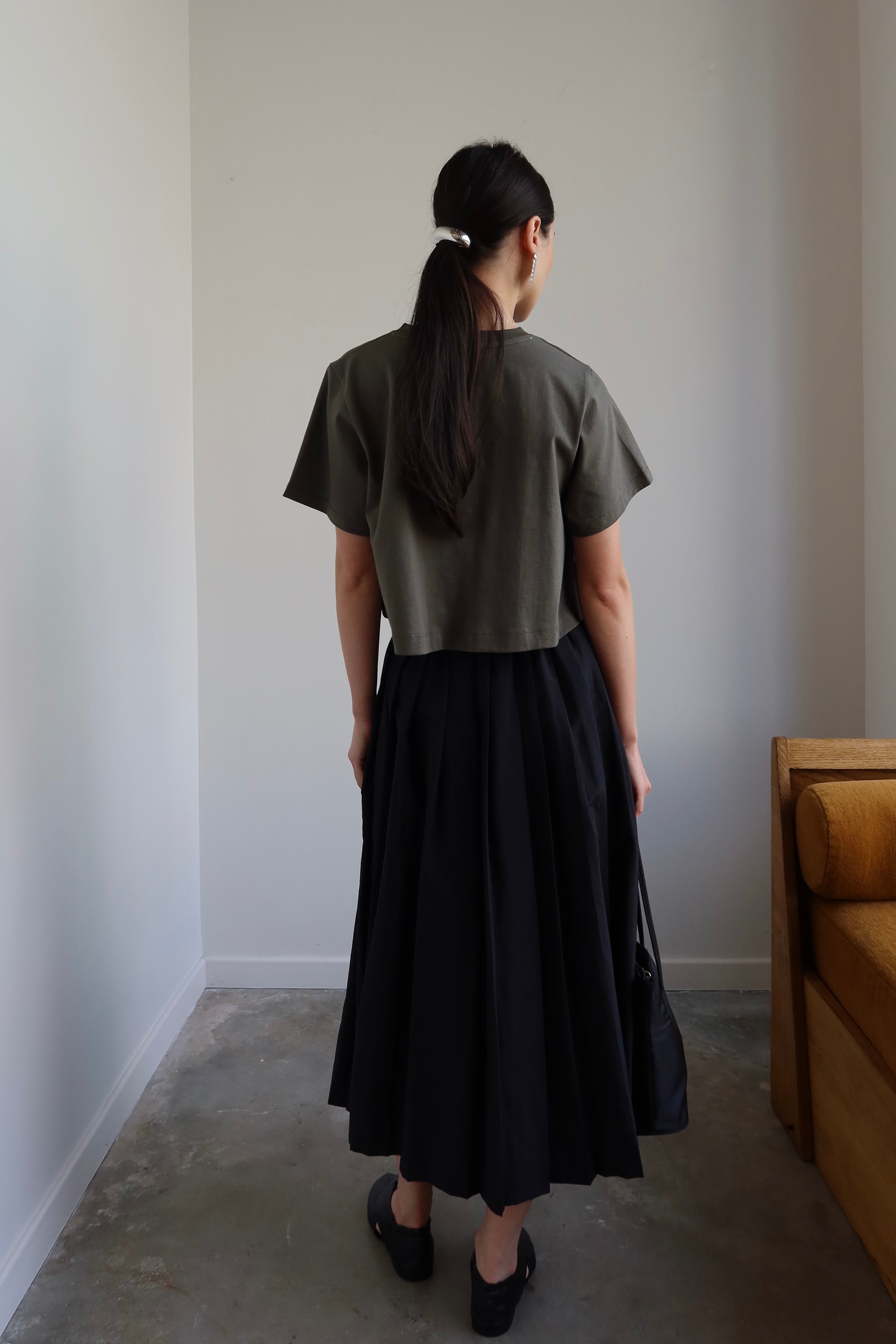 Back view of a woman standing looking over her shoulder wearing an Alas Eius black nylon pleated midi skirt paired with black leather flats and a cropped t-shirt at Alas Eius. 