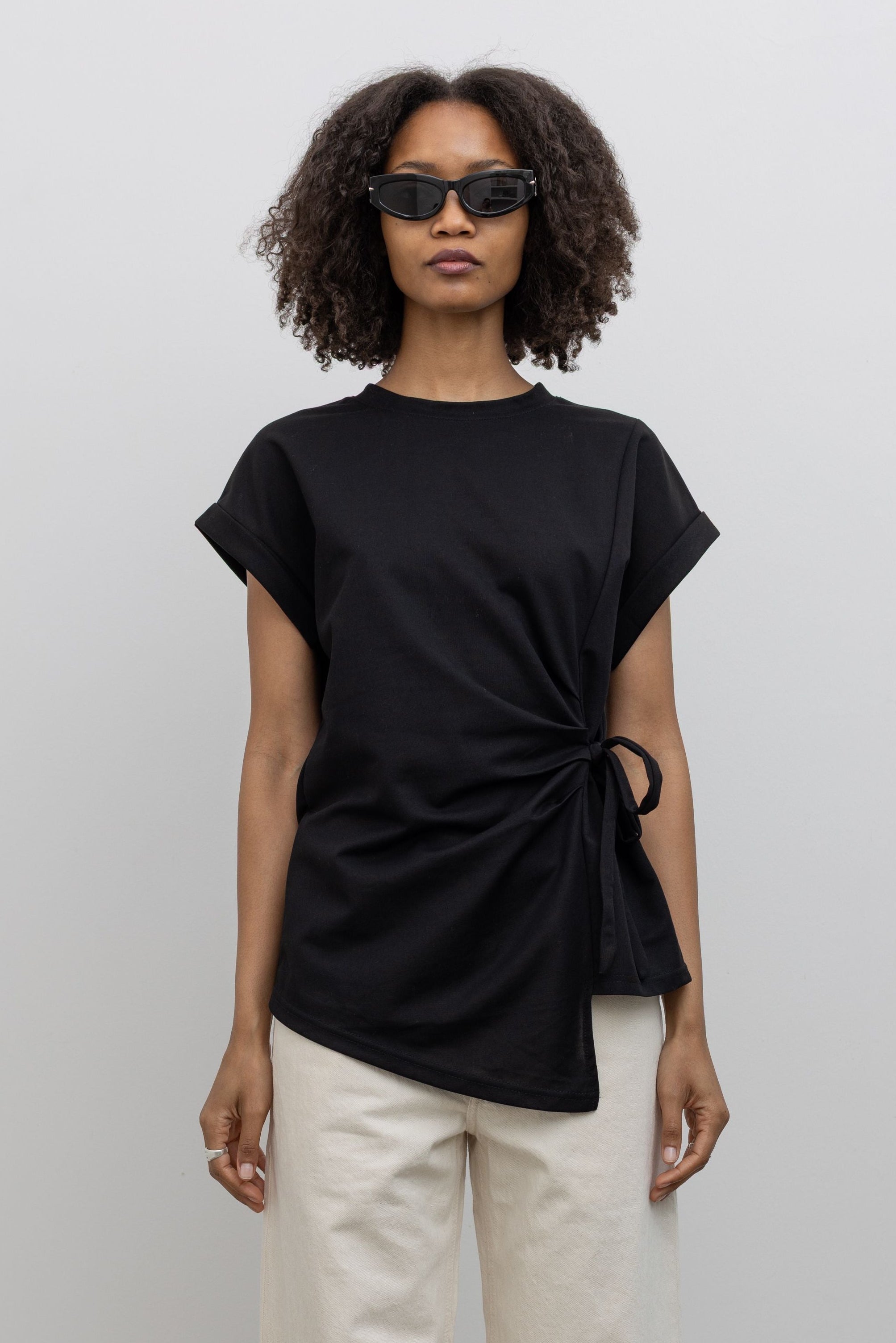Front view of a woman standing wearing an elevated take on the black standard tee in an Alas Eius label black cotton blend side tie asymmetrical hem short sleeve top with white denim jeans at an Alas Eius store. 