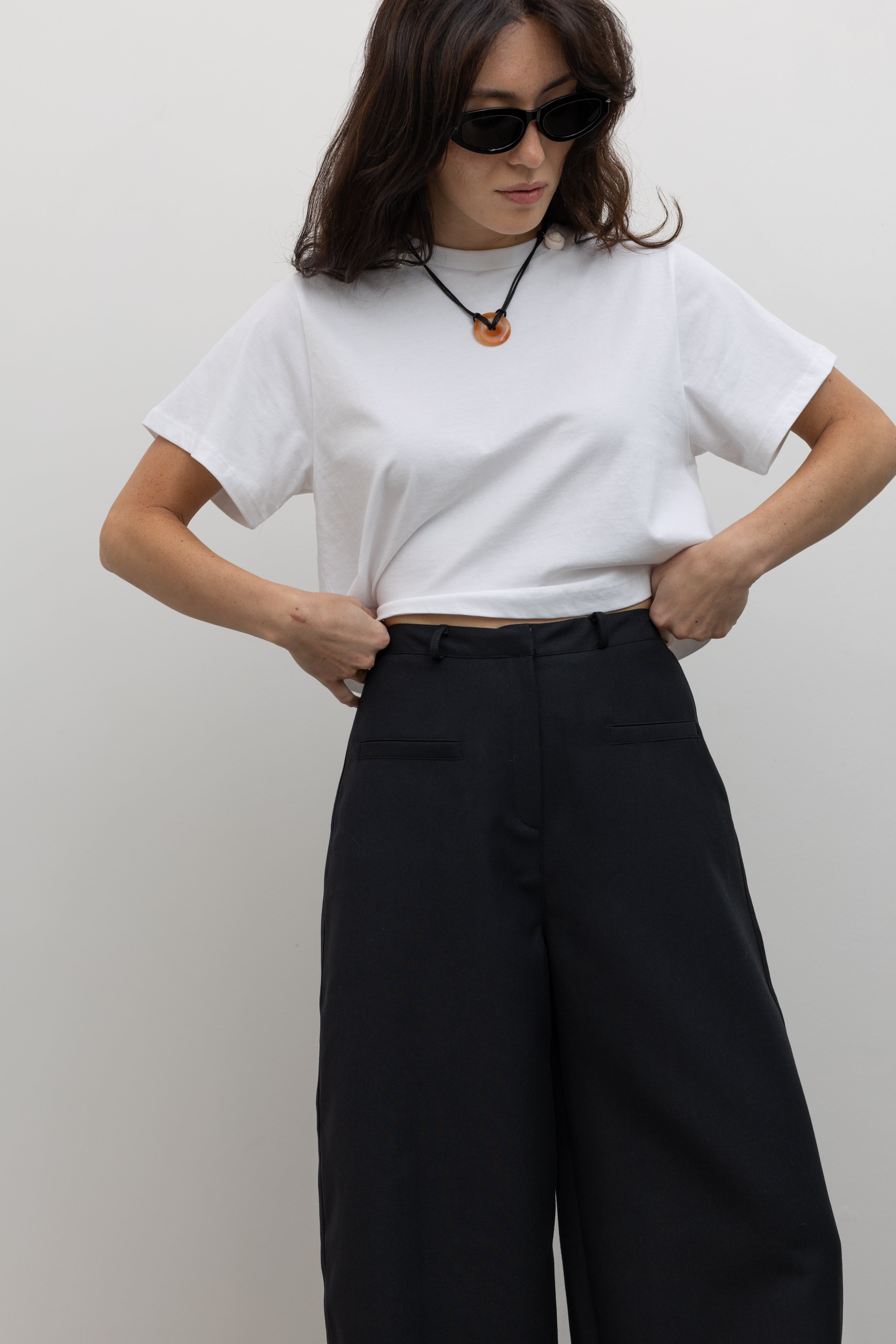 Close-up front view of a woman standing wearing minimalist fashion in a Alas Eius label black wool carrot high-waisted, wide-leg trouser paired with an Alas Eius label white cotton boxy crop top at Alas Eius. 