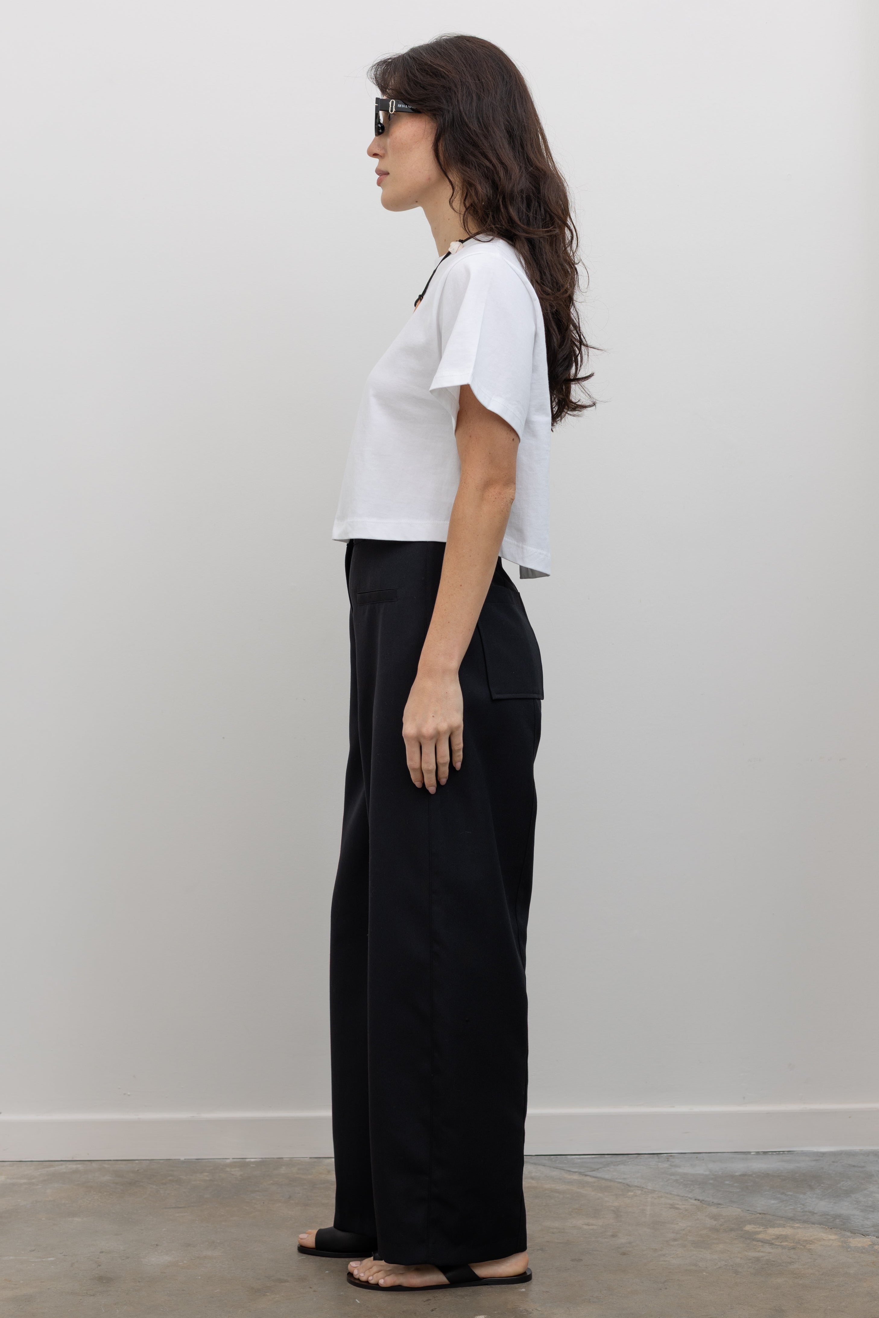 Full length side view of a woman standing wearing modern minimalist spring fashion in an Alas Eius black wool carrot high-waisted, wide-leg trouser paired with a white boxy cropped top at Alas Eius. 