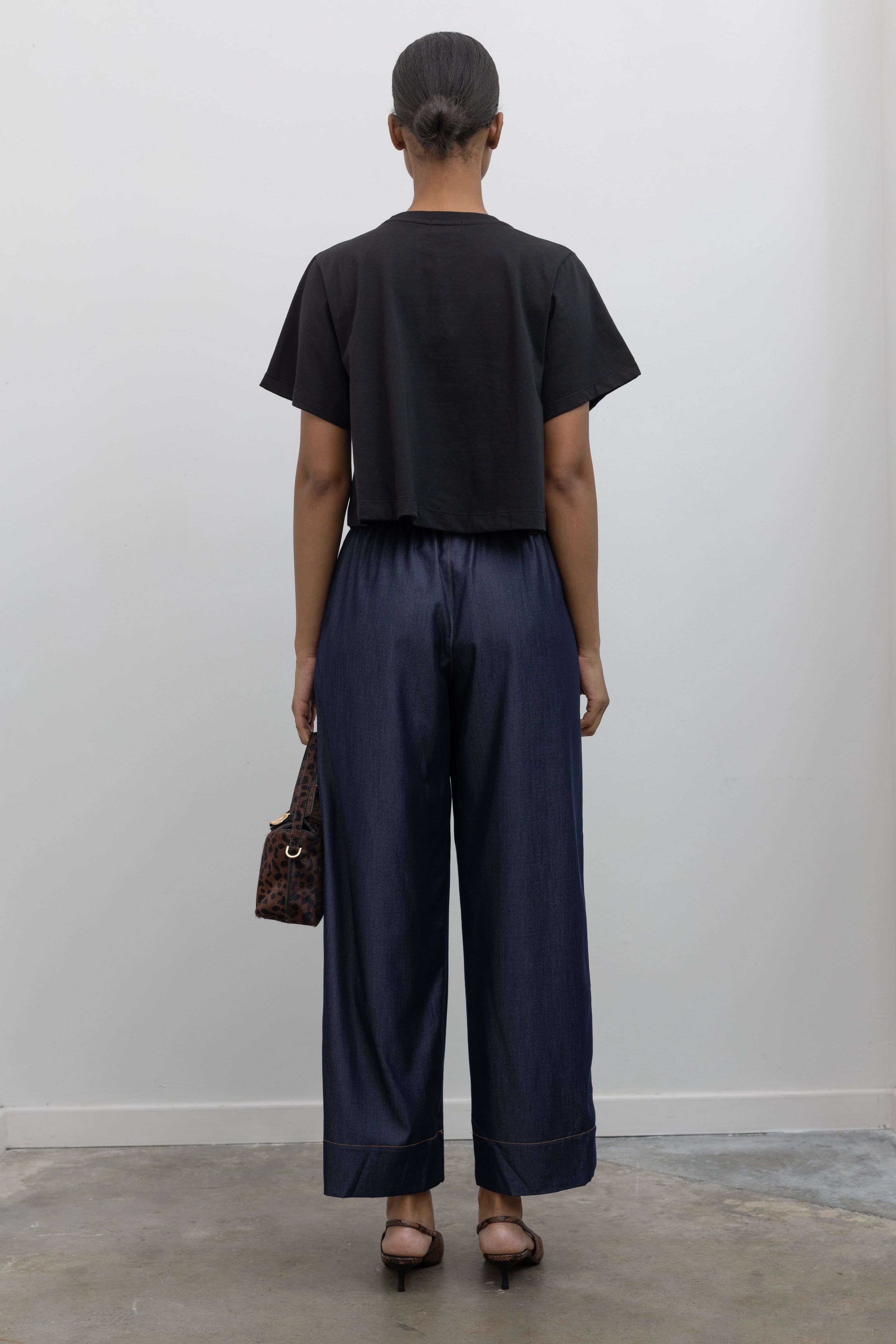 Back view of Woman standing wearing an everyday outfit with a black cotton short sleeved cropped t-shirt and Alas Eius Label Tencel Blend Dark Wash Tailored Wide Leg Pull On Pants with slingback kitten heels and accessories at Alas Eius. 