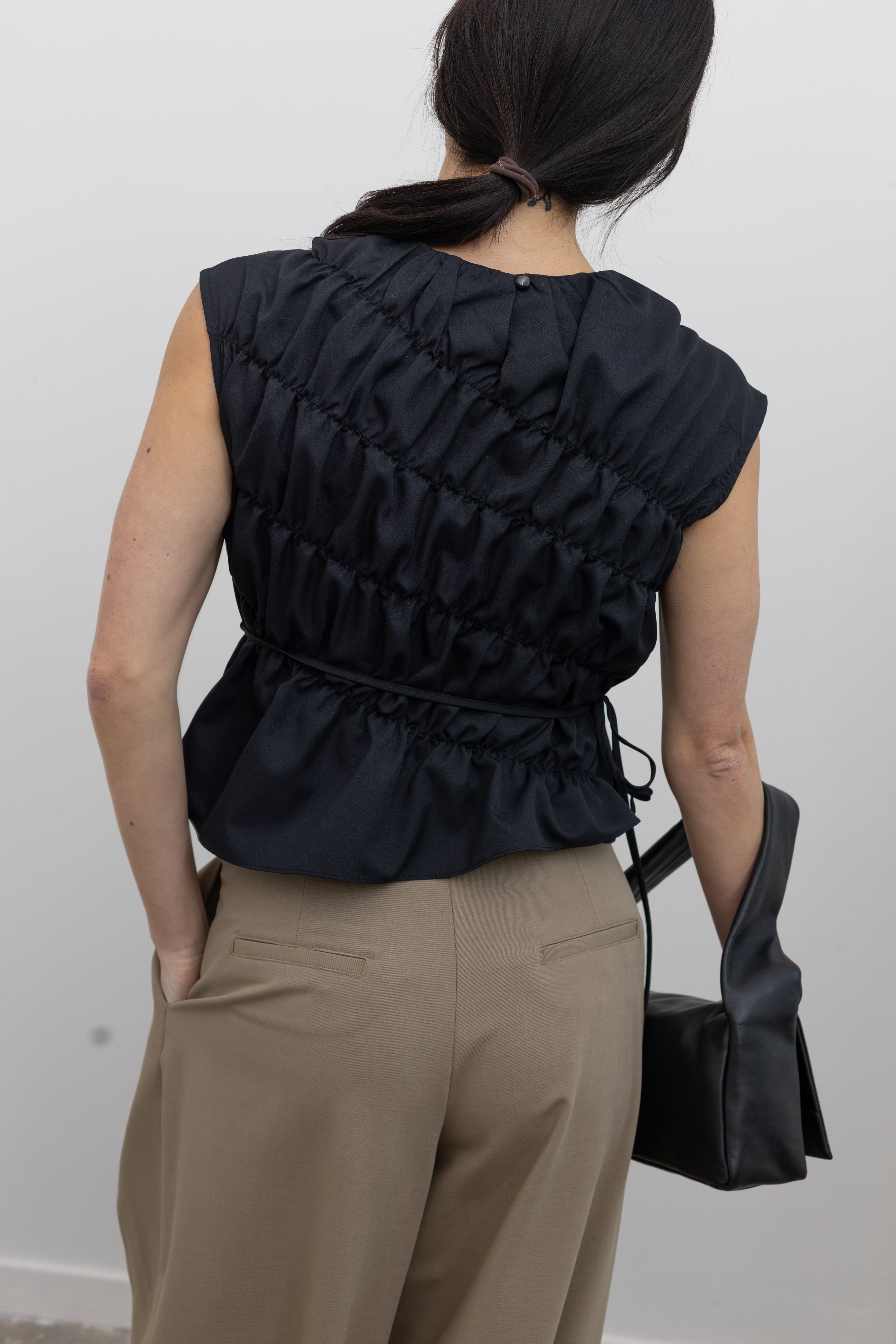 Close-up back view of a woman posing wearing contemporary and chic women's fashion in an Alas Eius Black Asymmetrical Cinched Top featuring a crew neckline, cap sleeves, and an optional waist tie paired with elevated khaki cinched ankle pants, silver sandals, and a black leather St. Agni bag at an Alas Eius store. 