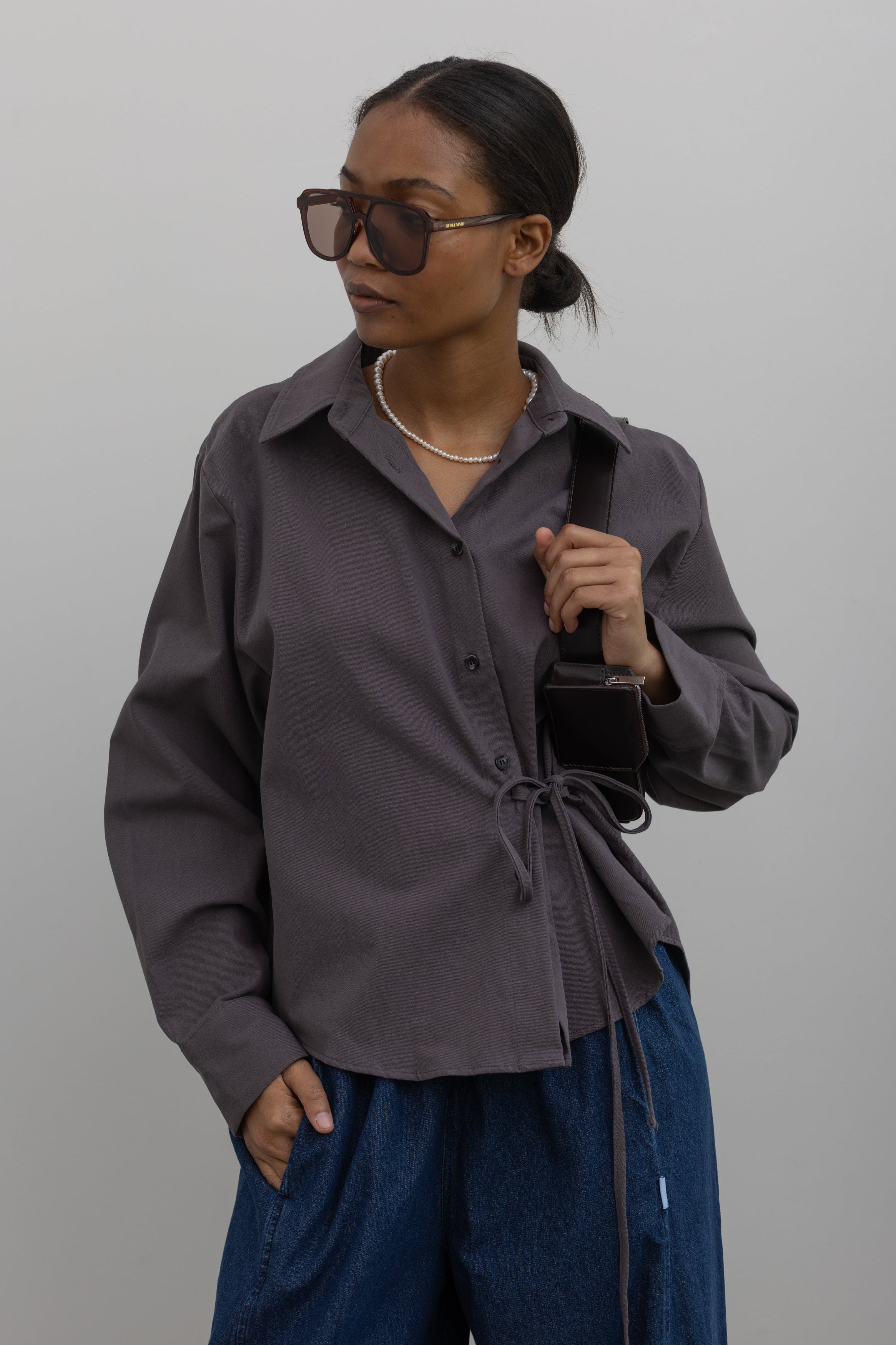 Woman wearing capsule wardrobe essential in a modern and structural Alas Eius label cinder gray brushed cotton cinched waist button down collared long sleeve top with denim and black leather handbag Alas Eius