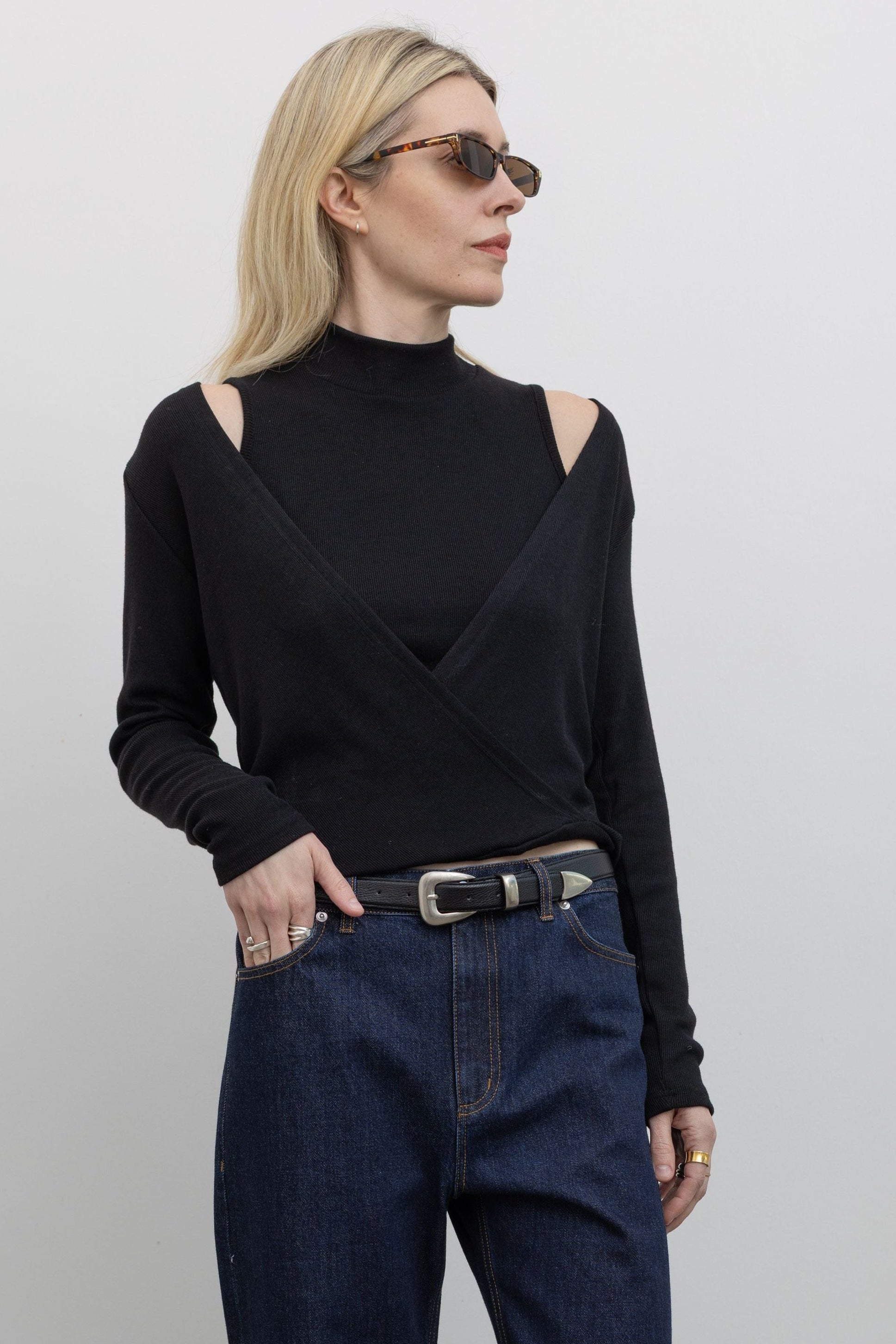 Woman wearing modern minimalist spring fashion in a Alas Eius label cold shoulder mock neck black lightweight sweater with distinct cutouts at shoulder and a deep v-neck line paired with dark wash denim Tibi jeans and a classic black belt at Alas Eius. 