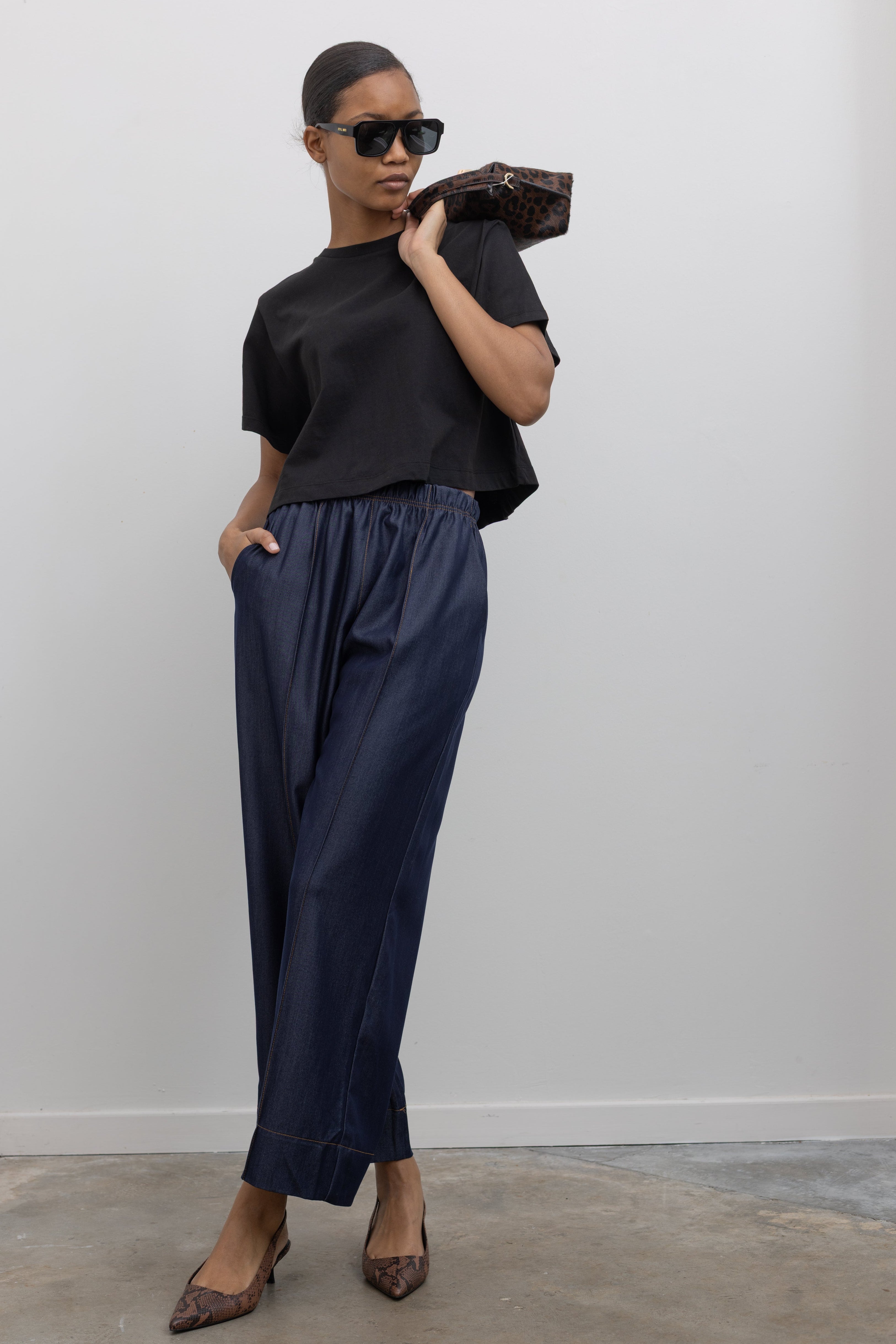 Woman standing wearing an everyday outfit with a black cotton short sleeved cropped t-shirt and Alas Eius Label Tencel Blend Dark Wash Tailored Wide Leg Pull On Pants with slingback kitten heels and accessories at Alas Eius. 