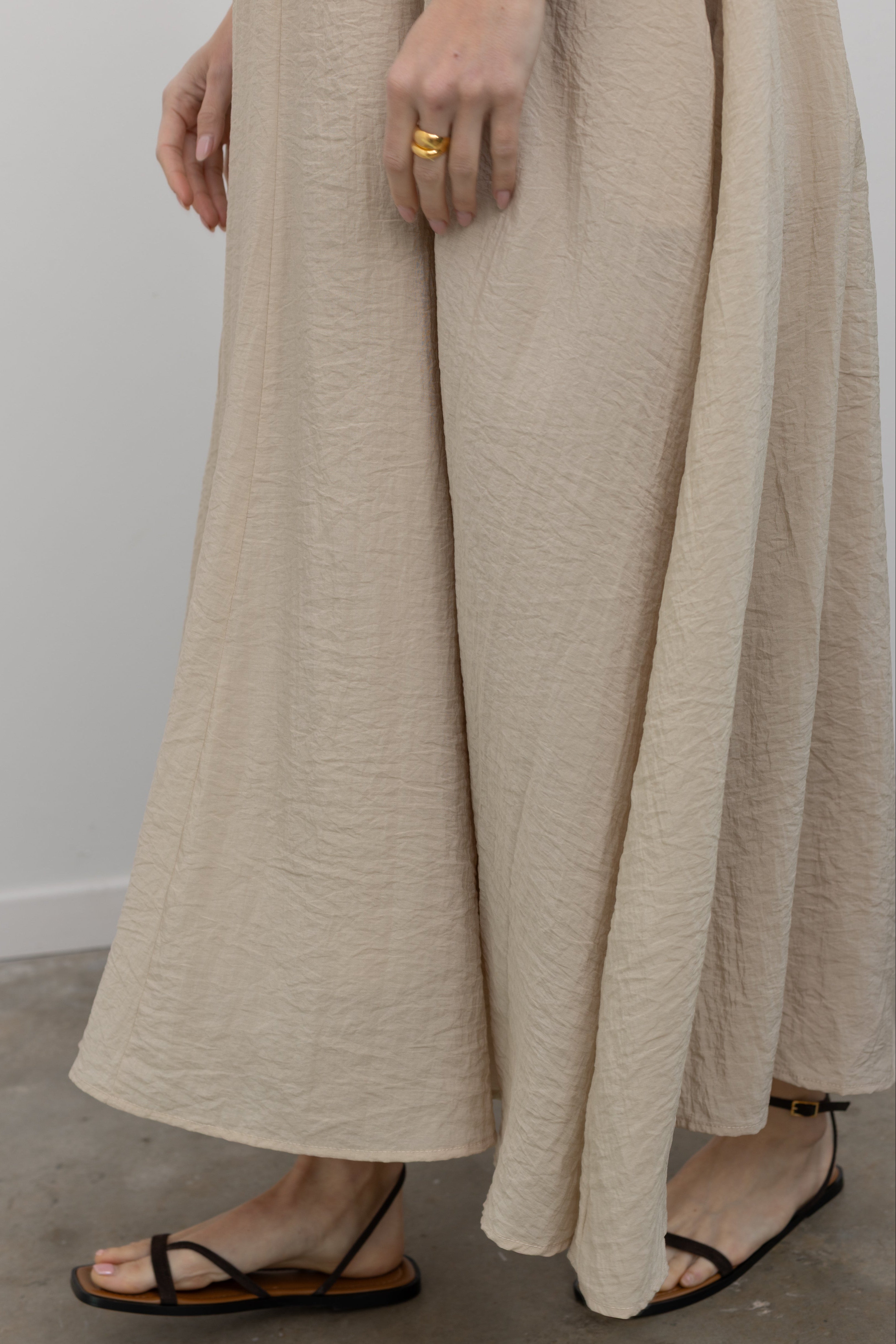 Close-up front view of a woman wearing an elegant and flowy versatile spring and summer dress in an Alas Eius label Beige V-Neck Thin Strap Dress in a beige cotton blend featuring a flowing silhouette, adjustable bungee straps for a customized fit and two convenient side pockets paired with thin strappy black leather sandals at an Alas Eius store. 