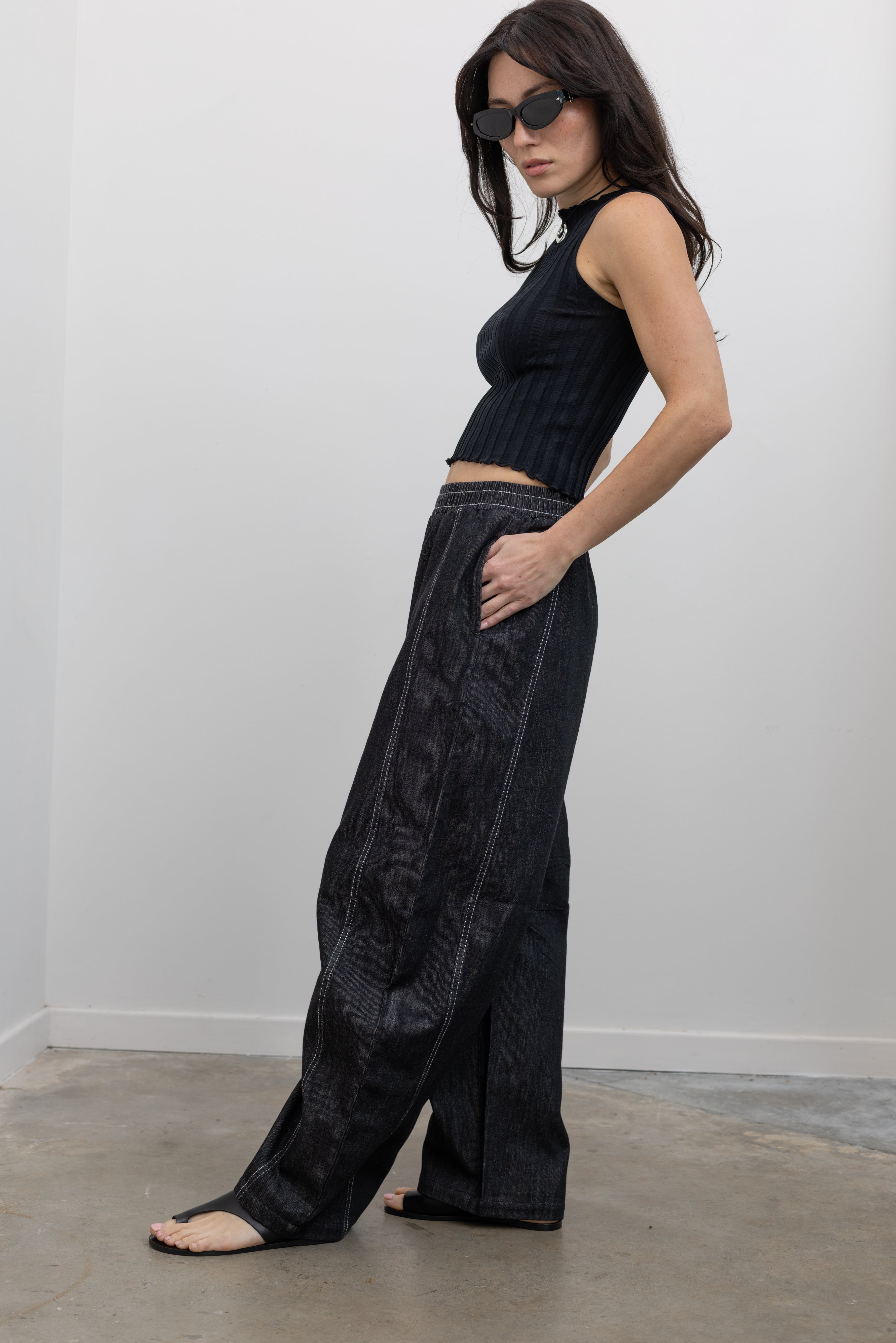 Full length side view of a woman standing wearing modern minimalist urban wear in the Alas Eius label black cotton barrel leg pants featuring contrasting white double stitching, side seam pockets, and an elastic waistband paired with a black ribbed knit crop tank and ATP black leather cut out sandals at an Alas Eius store. 