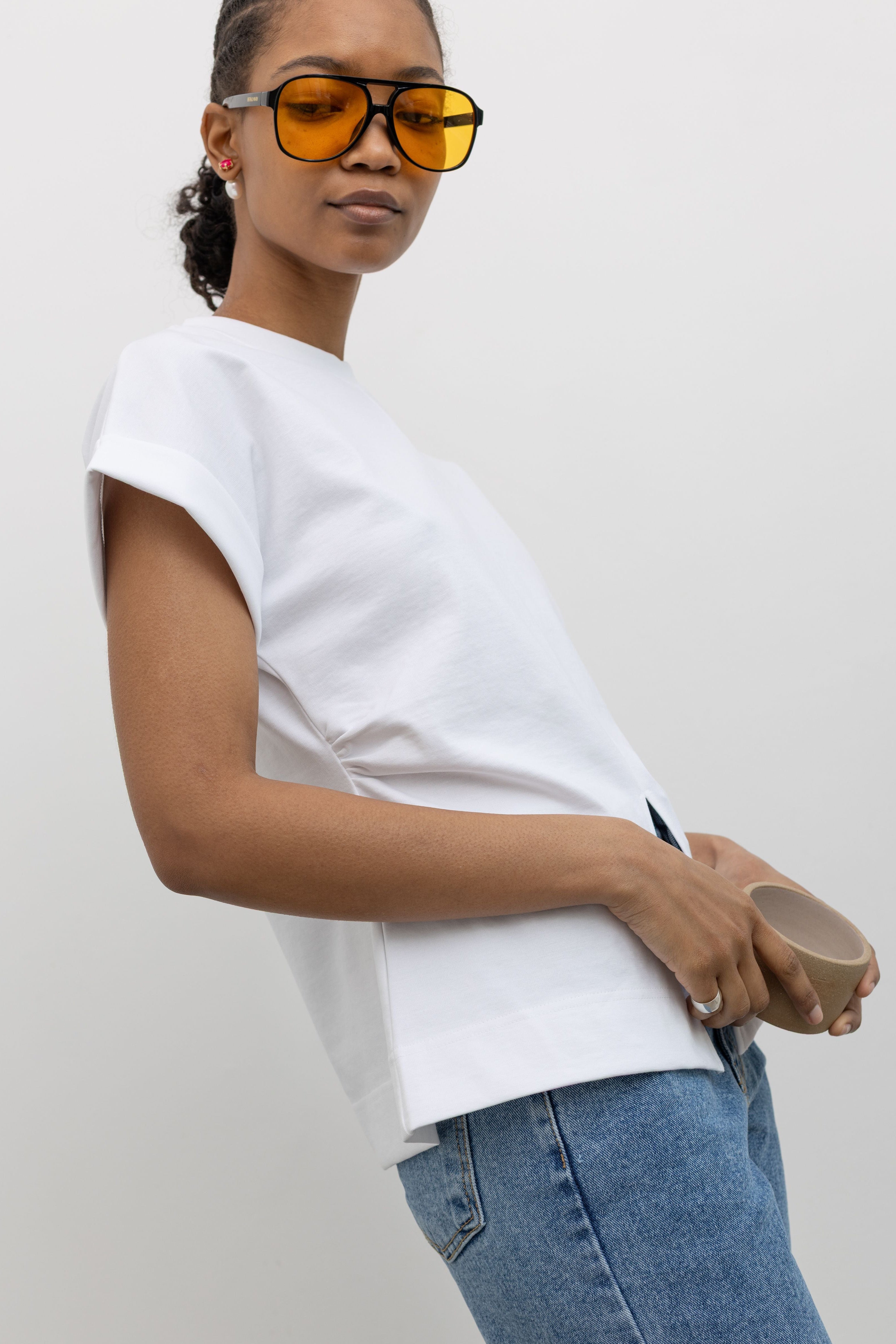 Side view of a woman posing with sunglasses wearing an elevated spring staple in a Alas Eius label white cinched waist rounded top designed stitched cuffed short sleeves, ruched side seams and an open v-cut vent at the front paired with light wash denim jeans at an Alas Eius store. 