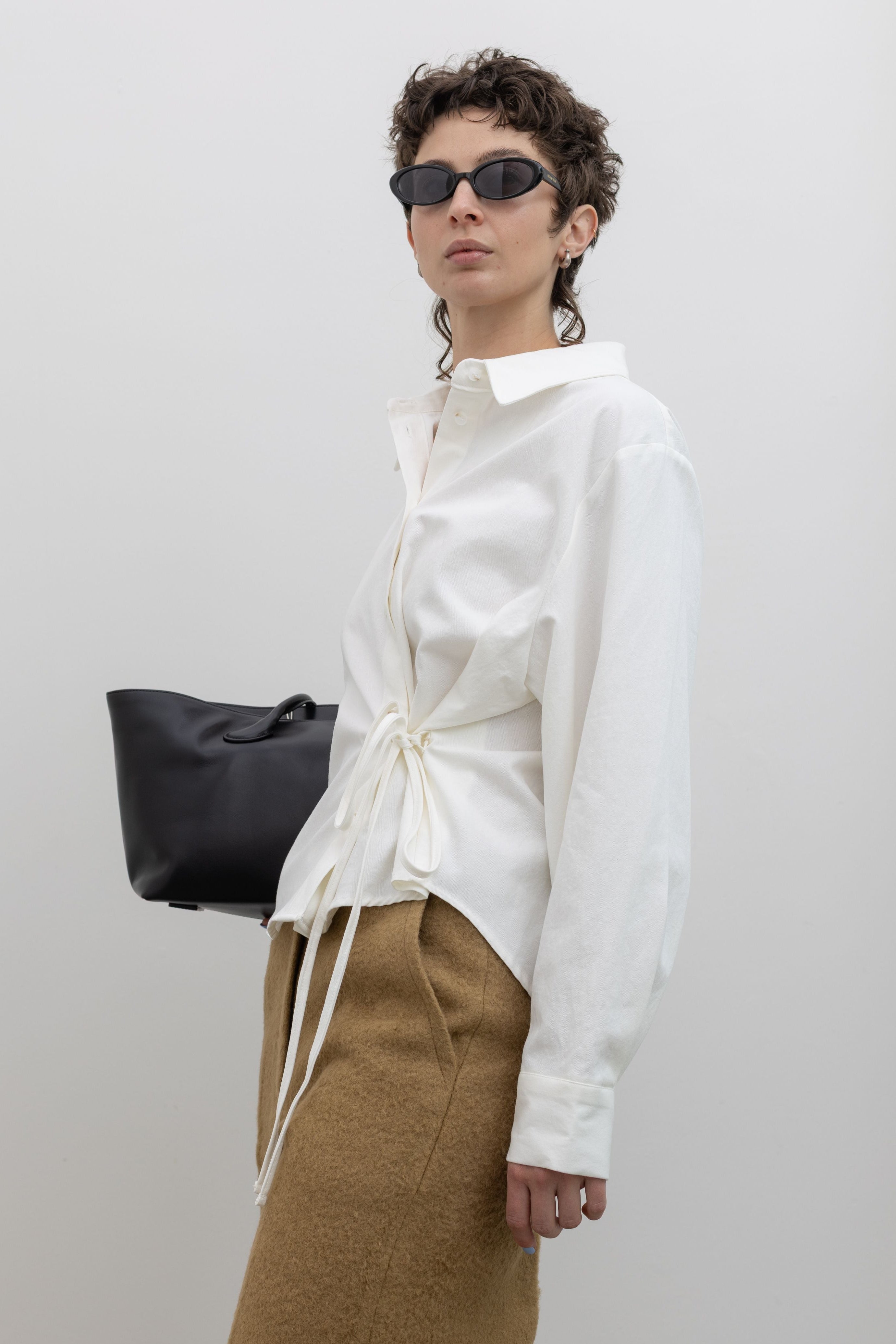 Three quarters view of a woman wearing capsule wardrobe essential style in an elegant and flattering Alas Eius label white brushed cotton side-cinched waist tie detail button down top paired with tan trousers and accessories at Alas Eius.