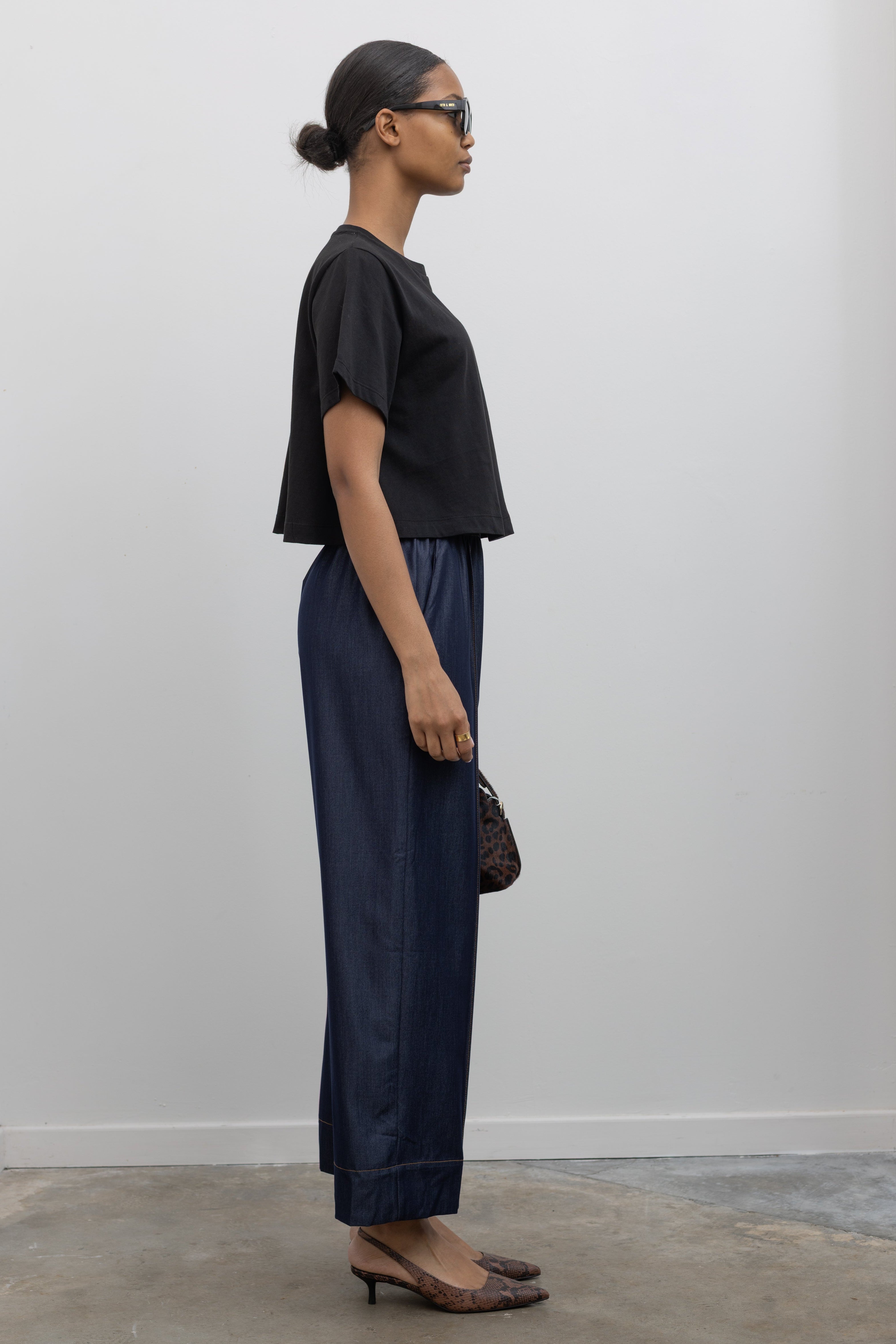 Side view of Woman standing wearing an everyday outfit with a black cotton short sleeved cropped t-shirt and Alas Eius Label Tencel Blend Dark Wash Tailored Wide Leg Pull On Pants with slingback kitten heels and accessories at Alas Eius. 