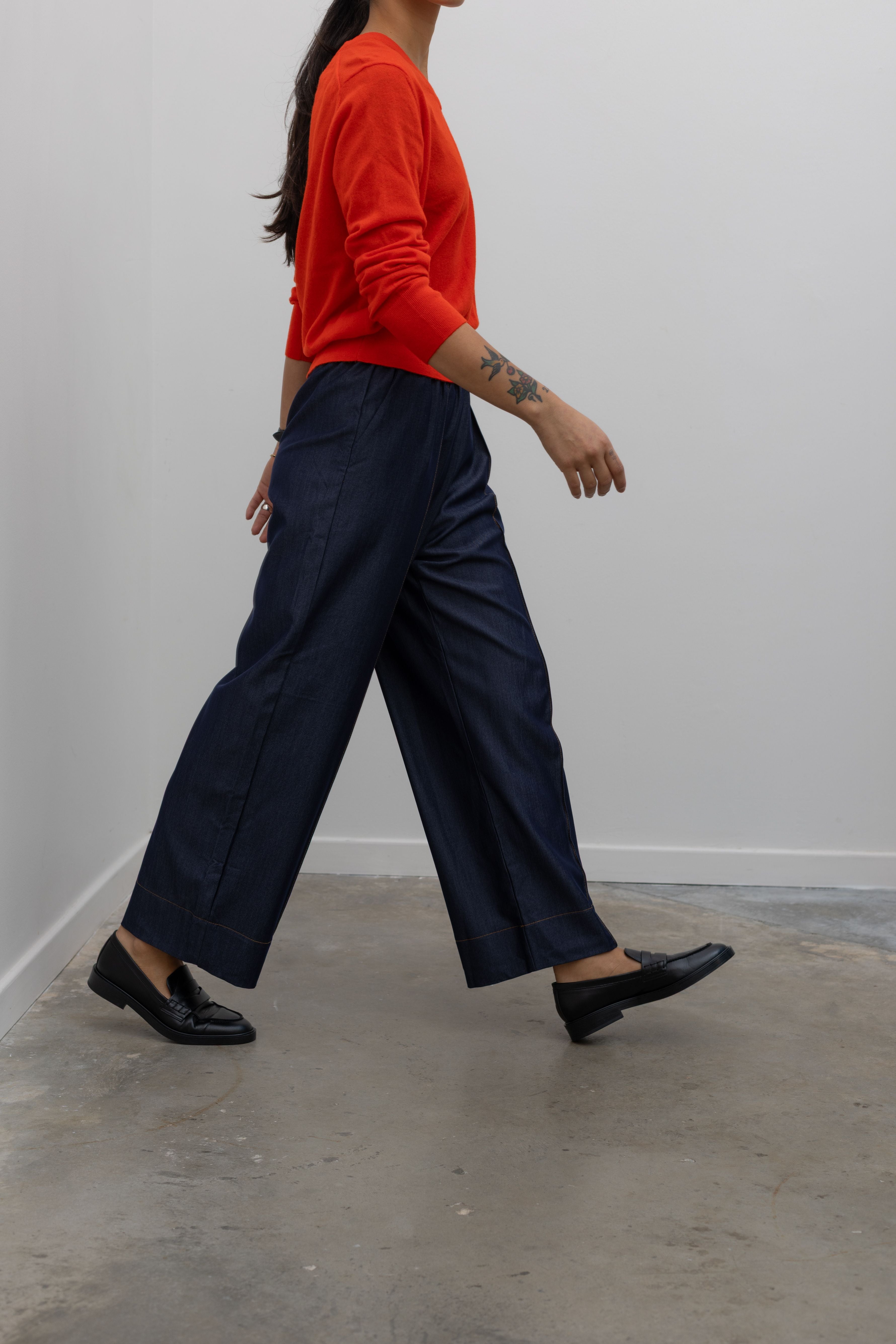 Side view of Woman in motion wearing an everyday outfit with a Tibi Red v-neck cashmere sweater and Alas Eius Label Tencel Blend Dark Wash Tailored Wide Leg Pull On Pants with black leather loafer at Alas Eius. 