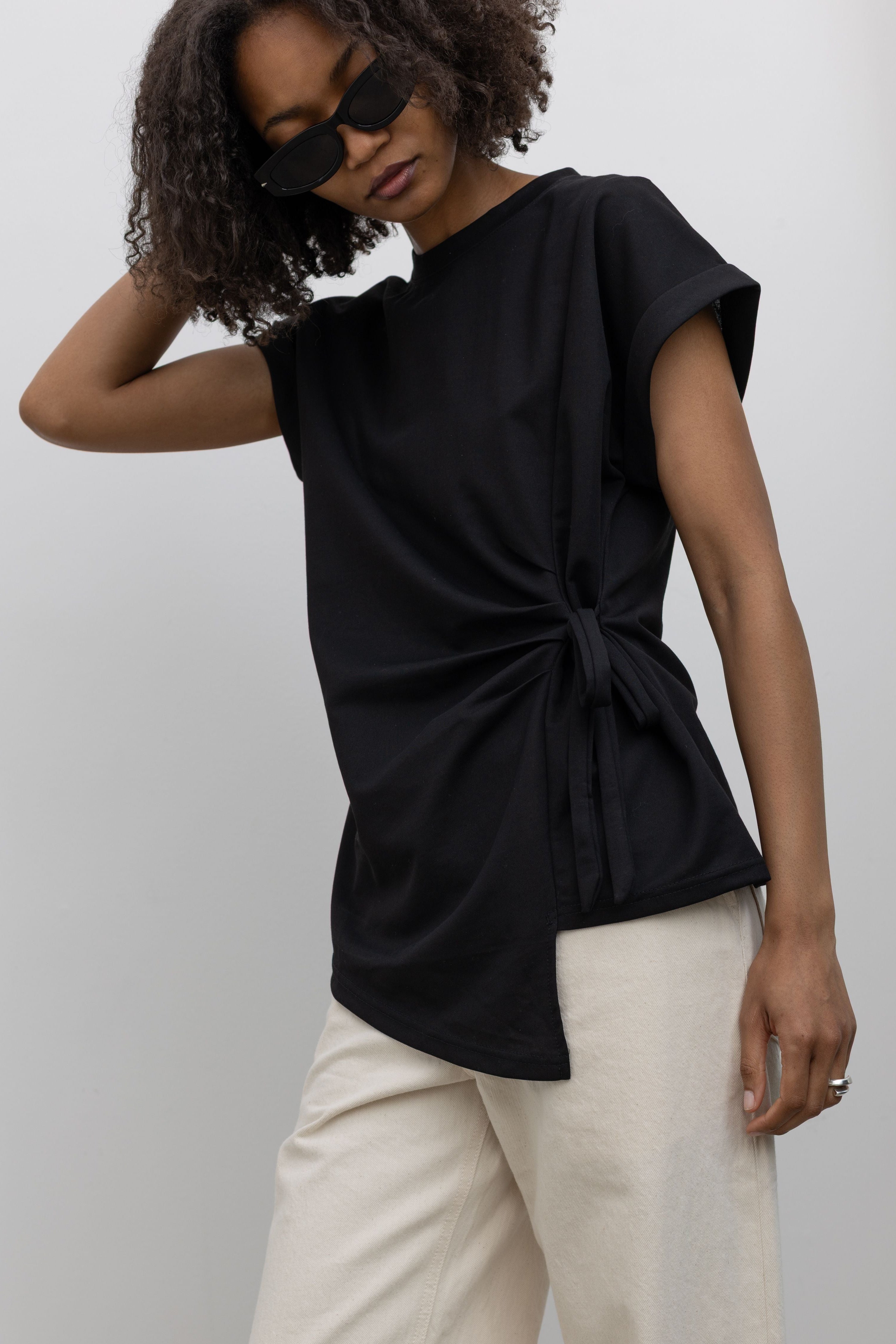 Front 3/4 view of a woman standing wearing an elevated take on the black standard tee in an Alas Eius label black cotton blend side tie asymmetrical hem short sleeve top with white denim jeans at an Alas Eius store. 