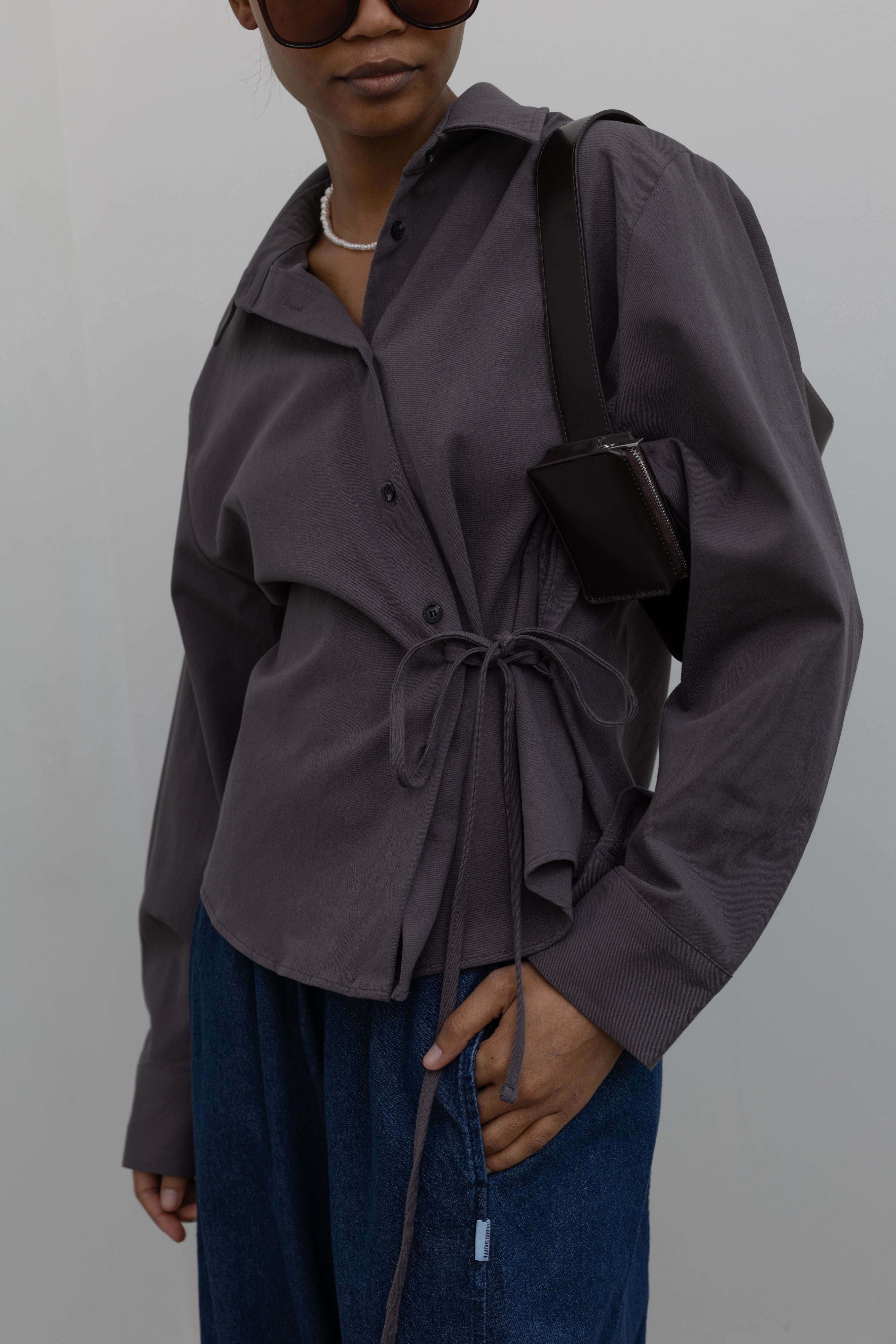 Detailed product view of Woman wearing capsule wardrobe essential in an Alas Eius label cinder gray brushed cotton tie waist button down collared long sleeve top with denim and a black leather handbag Alas Eius