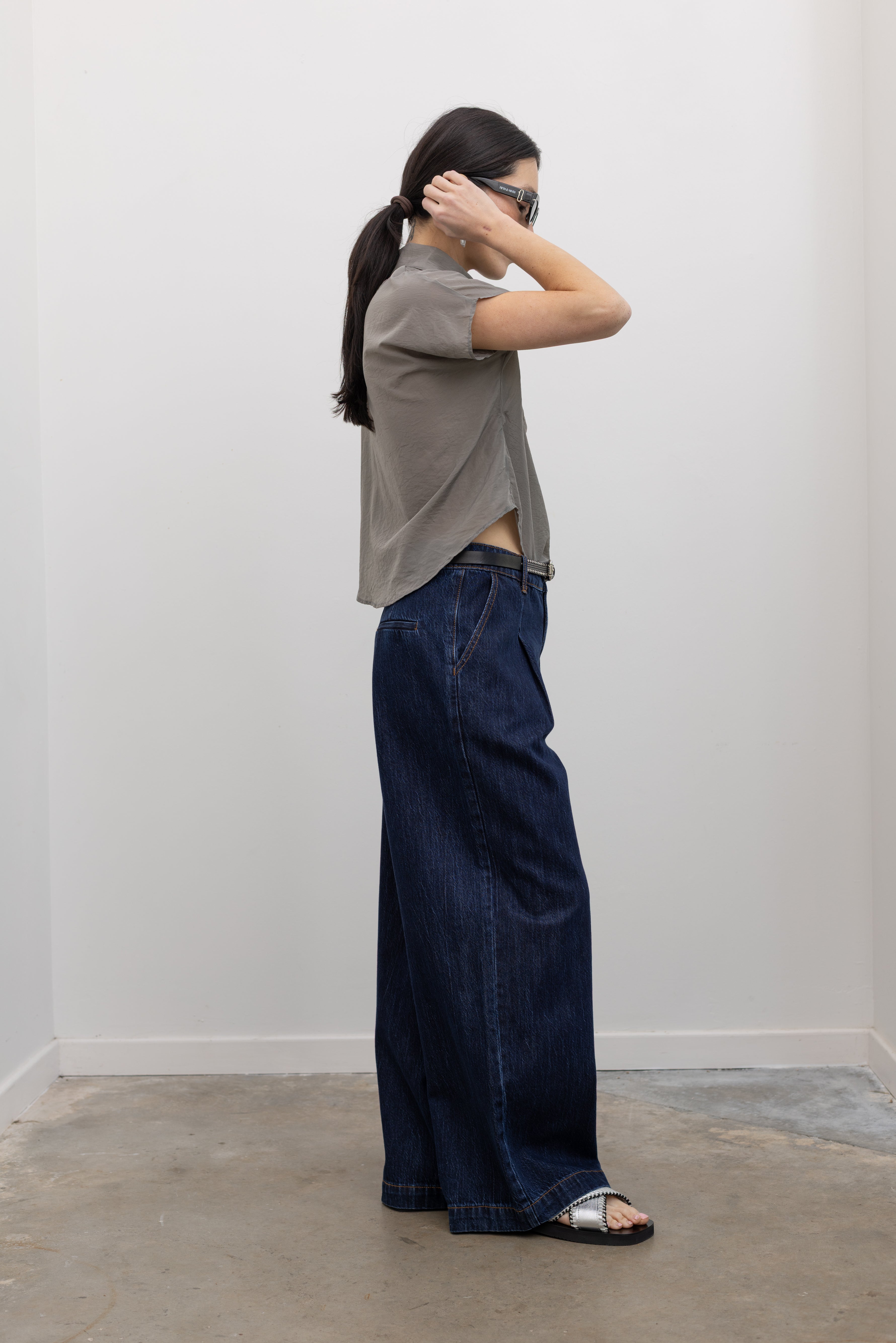 Full length side view of a woman wearing contemporary spring fashion in an Alas Eius label light 
gray organza asymmetrical button mock neck top with dark wash denim wide leg denim jeans at an Alas Eius store. 