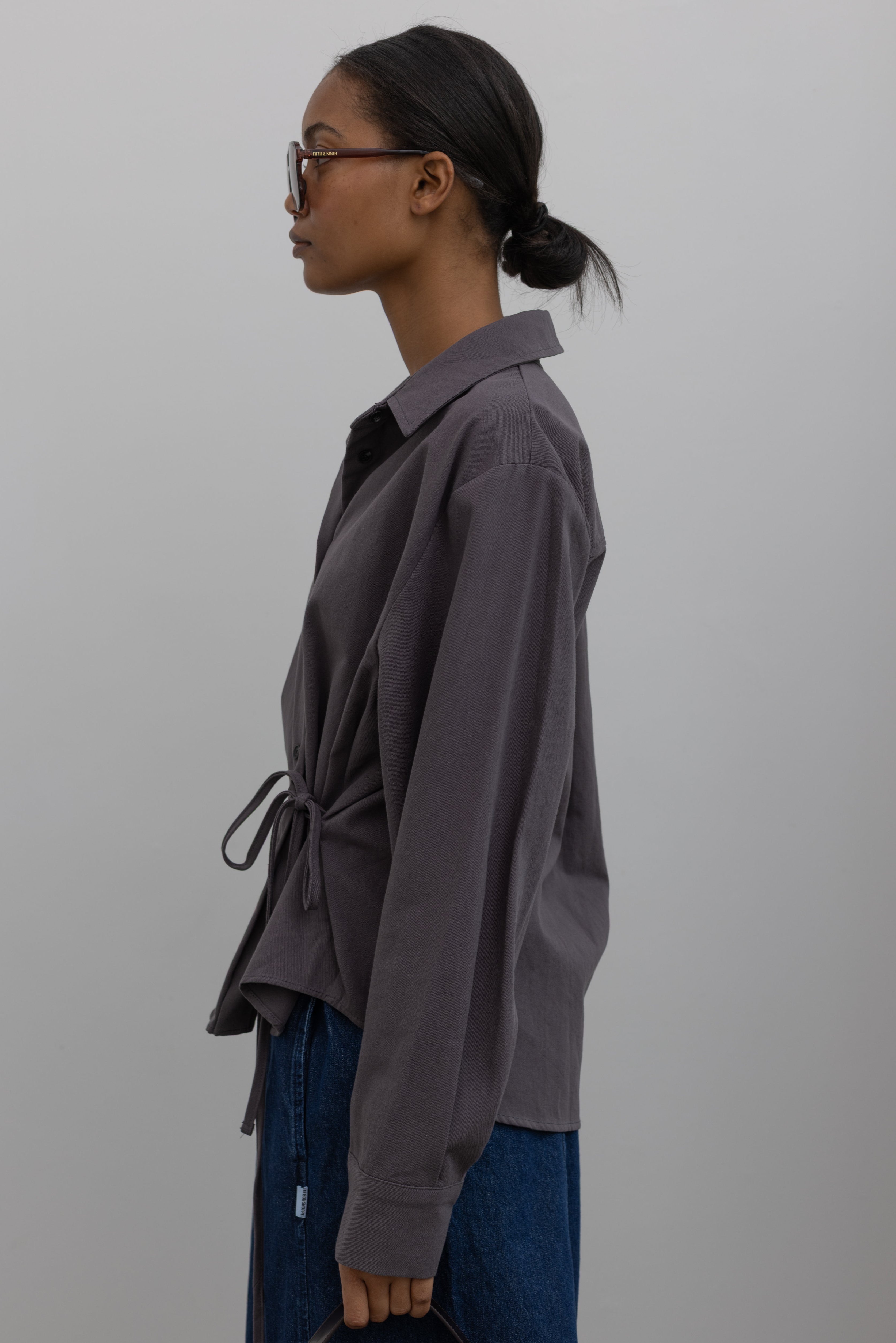 Left side view of Woman wearing capsule wardrobe essential in an elegant and flattering Alas Eius label gray brushed cotton cinched waist button down collared long sleeve top with denim at Alas Eius