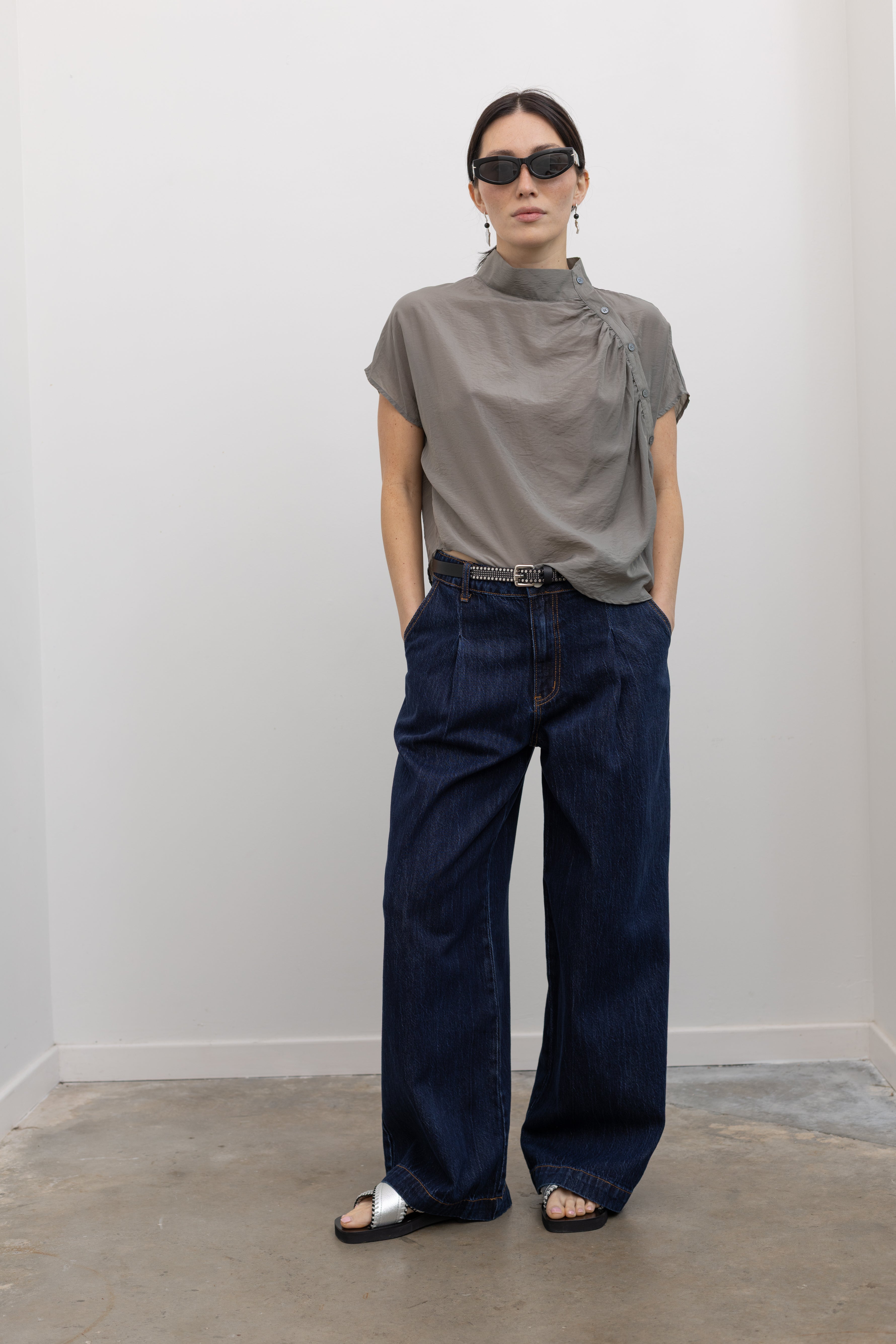 Full length front view of a woman wearing chic and elevated urban wear in an Alas Eius label light 
gray organza asymmetrical button mock neck top with dark wash denim jeans at an Alas Eius store. 
