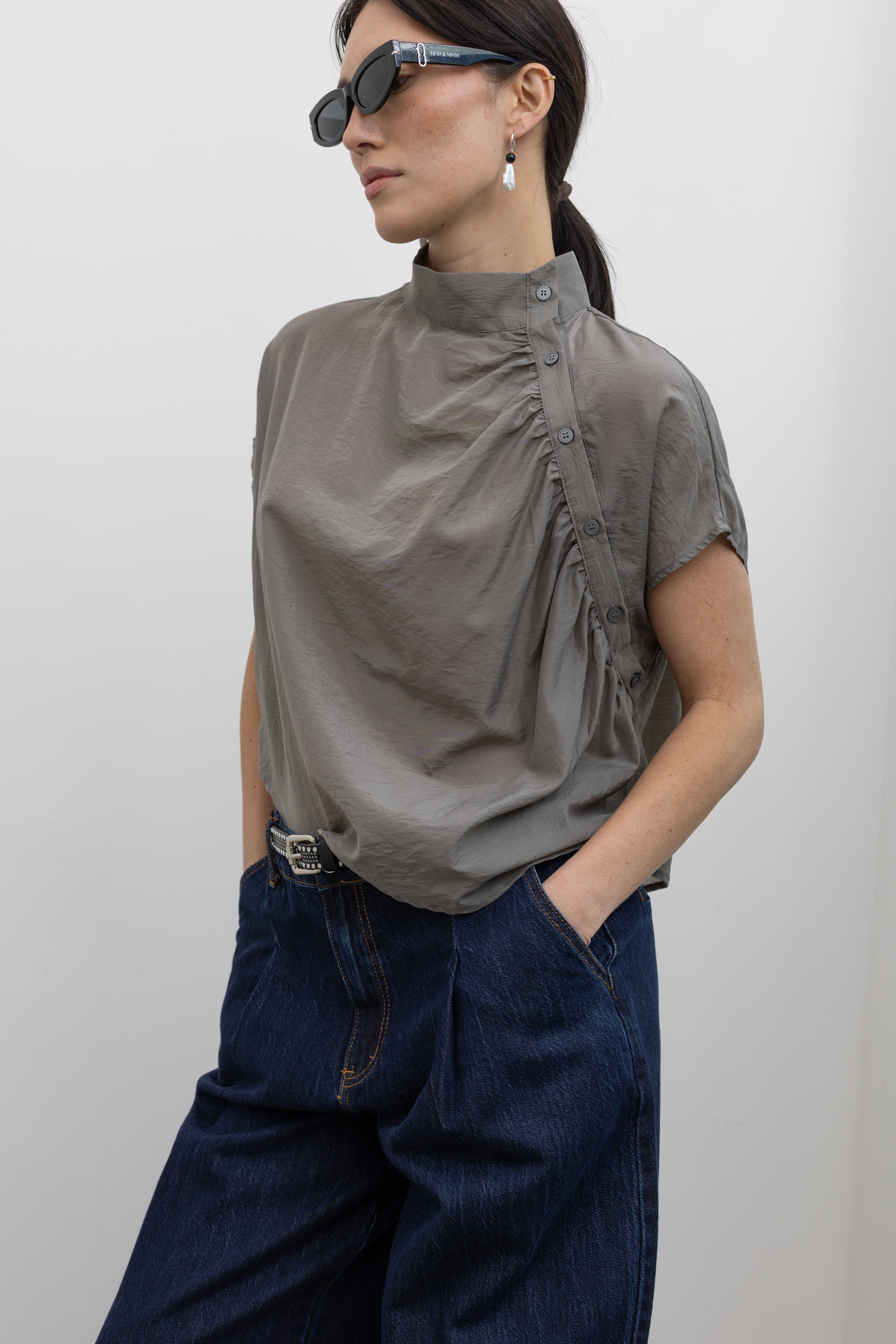 Close-up front view of a woman wearing modern minimalist spring fashion in an Alas Eius label light 
gray organza diagonal button front mock neck top with dark wash denim jeans at an Alas Eius store. 