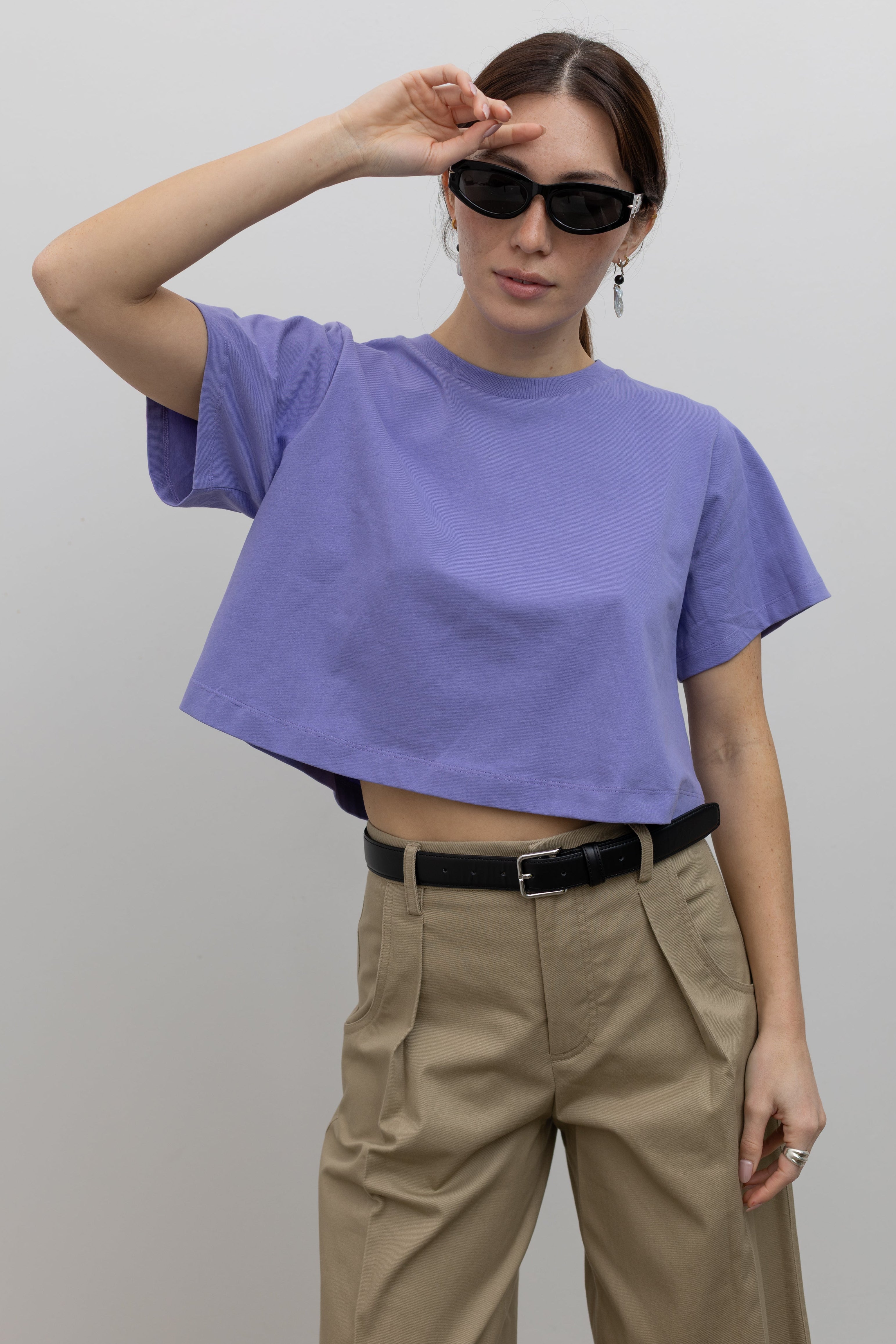Woman standing wearing must-have capsule wardrobe essential in an Alas Eius Label Lavender Relaxed Cropped T-Shirt with khaki pleated high waisted trousers and thin black leather belt with silver hardware. 