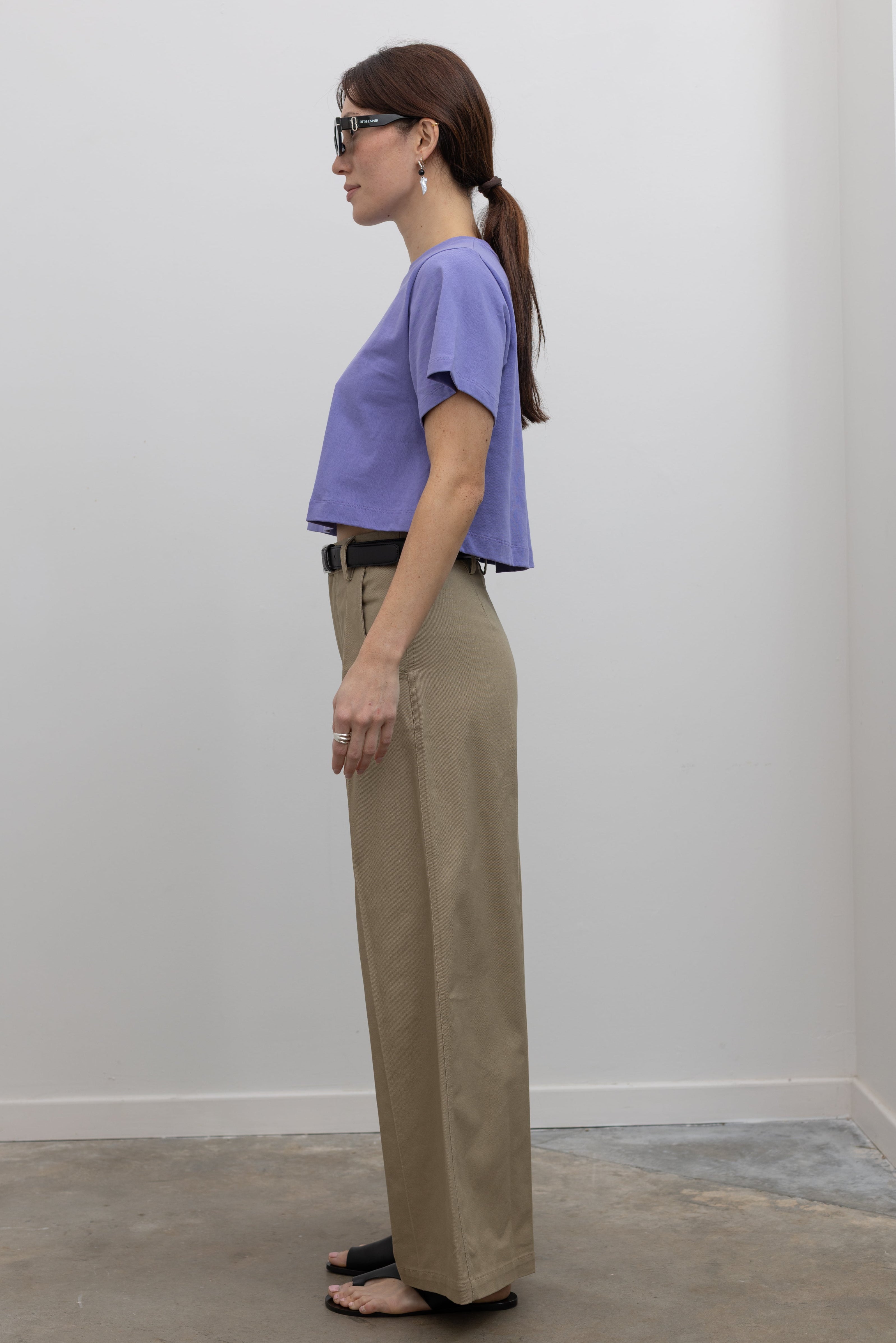 Side view of Woman standing wearing must-have capsule wardrobe essential in an Alas Eius Label Lavender Relaxed Boxy Cropped T-Shirt with khaki pleated high waisted trousers and thin black leather belt with silver hardware and black leather cutout sandals at Alas Eius 