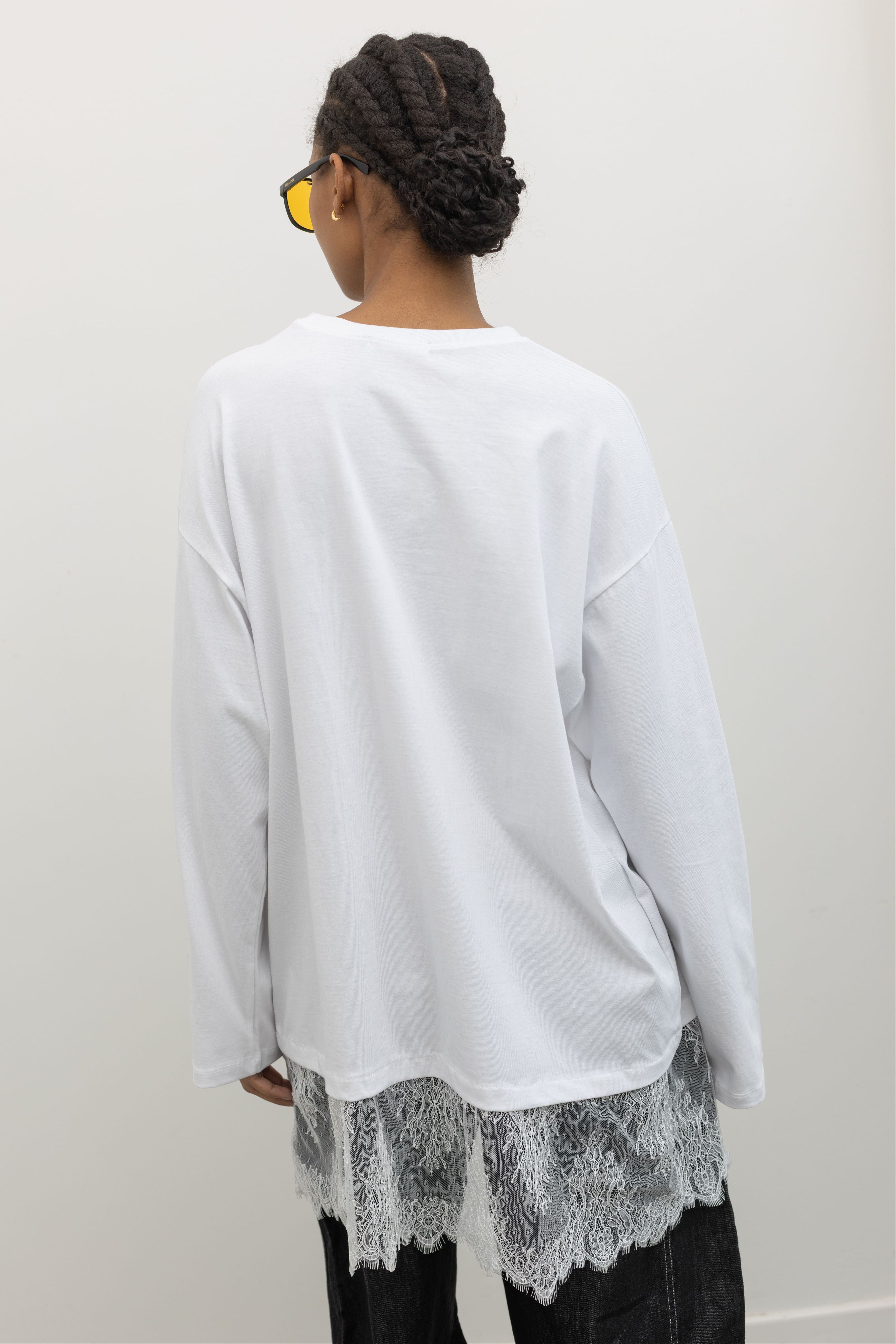 Close-up back view of a woman standing wearing spring fashion in a 
versatile Alas Eius label white long-sleeve oversized tunic top with lace hemline at and Alas Eius store. 