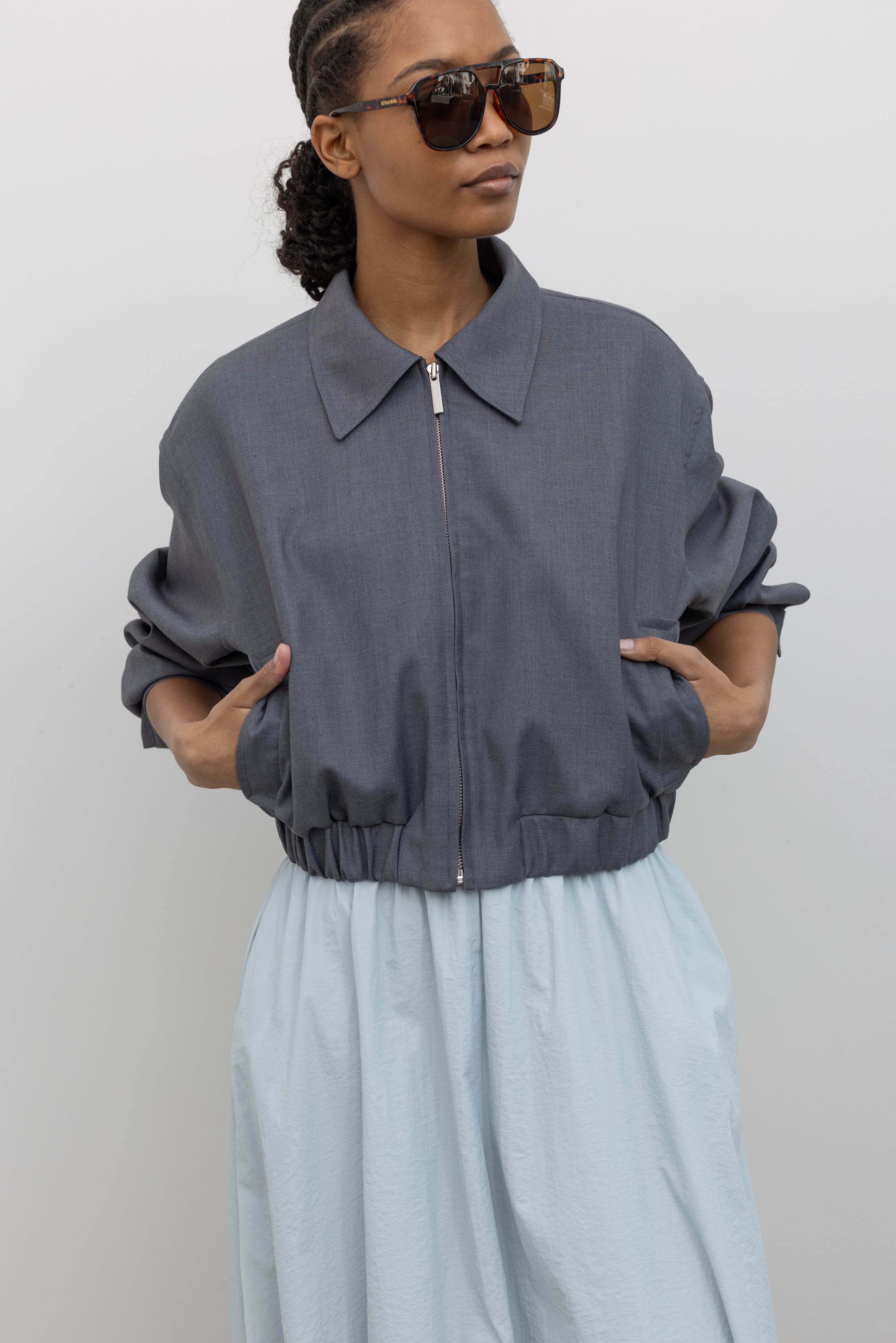 Front view of a woman wearing contemporary womenswear spring fashion in an Alas Eius label charcoal gray cropped tailored zip jacket featuring an elastic hem for a flattering cinched waist at an Alas Eius store. 