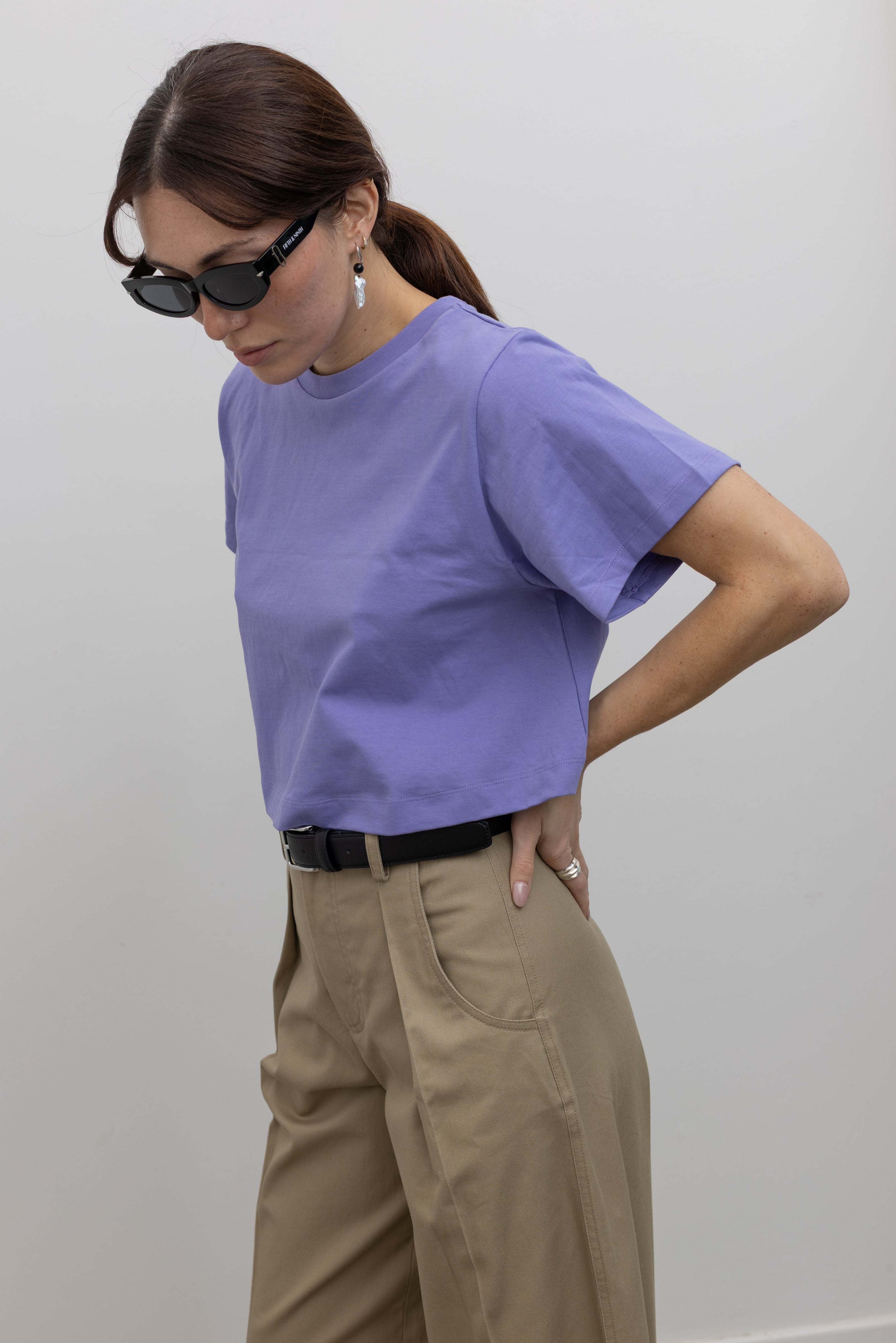 Detailed product view of a Woman standing wearing must-have capsule wardrobe essential in an Alas Eius Label Lavender Relaxed Boxy Cropped T-Shirt with khaki pleated high waisted trousers and thin black leather belt with silver hardware at Alas Eius 
