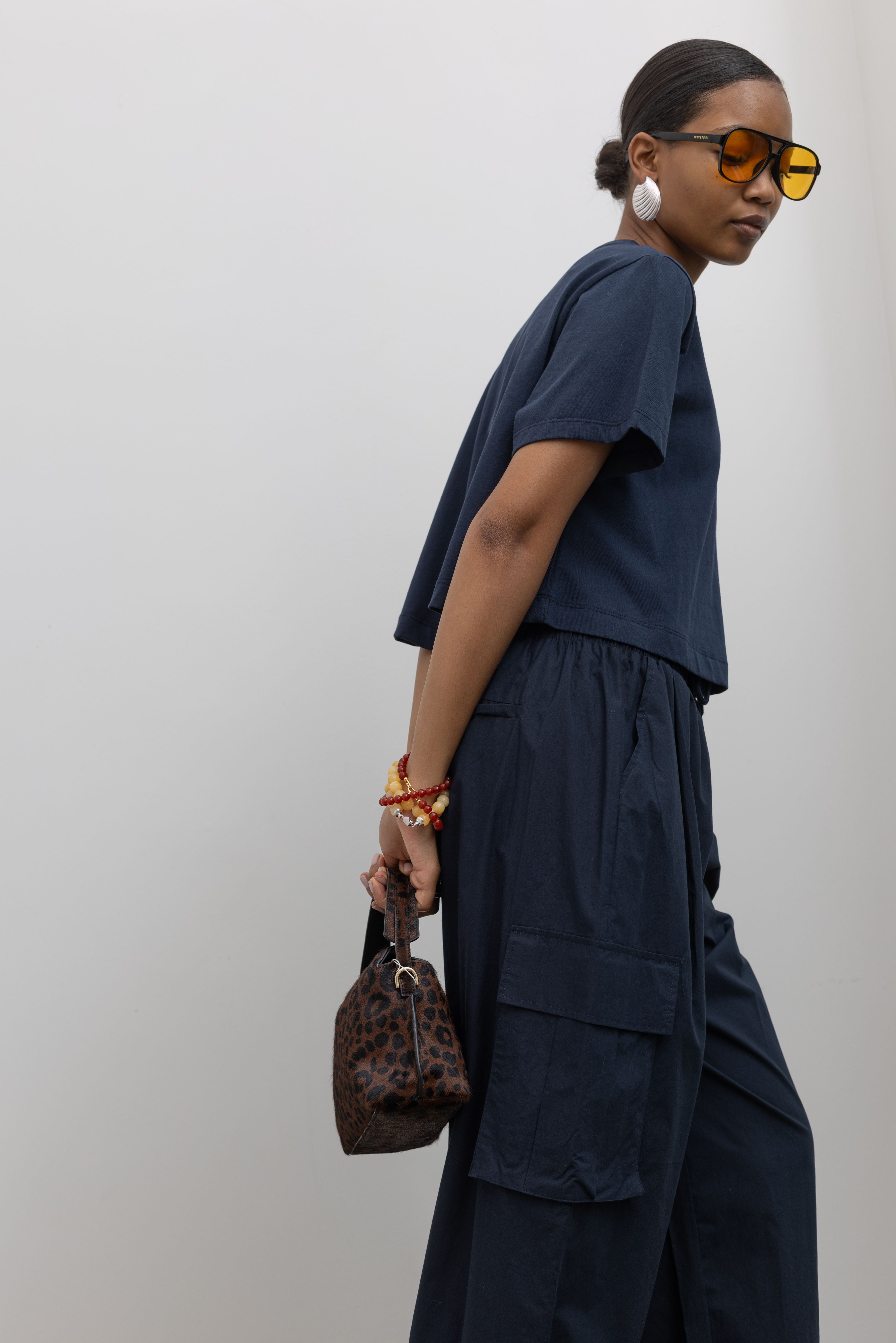 Side view of a Woman standing with arms clasped behind back wearing a capsule wardrobe essential in a pair of
Alas Eius navy blue lightweight cotton wide leg cargo pants. Featuring an oversized fit with drawstring ankles, a tie waist, and extra pockets on the legs at Alas Eius.