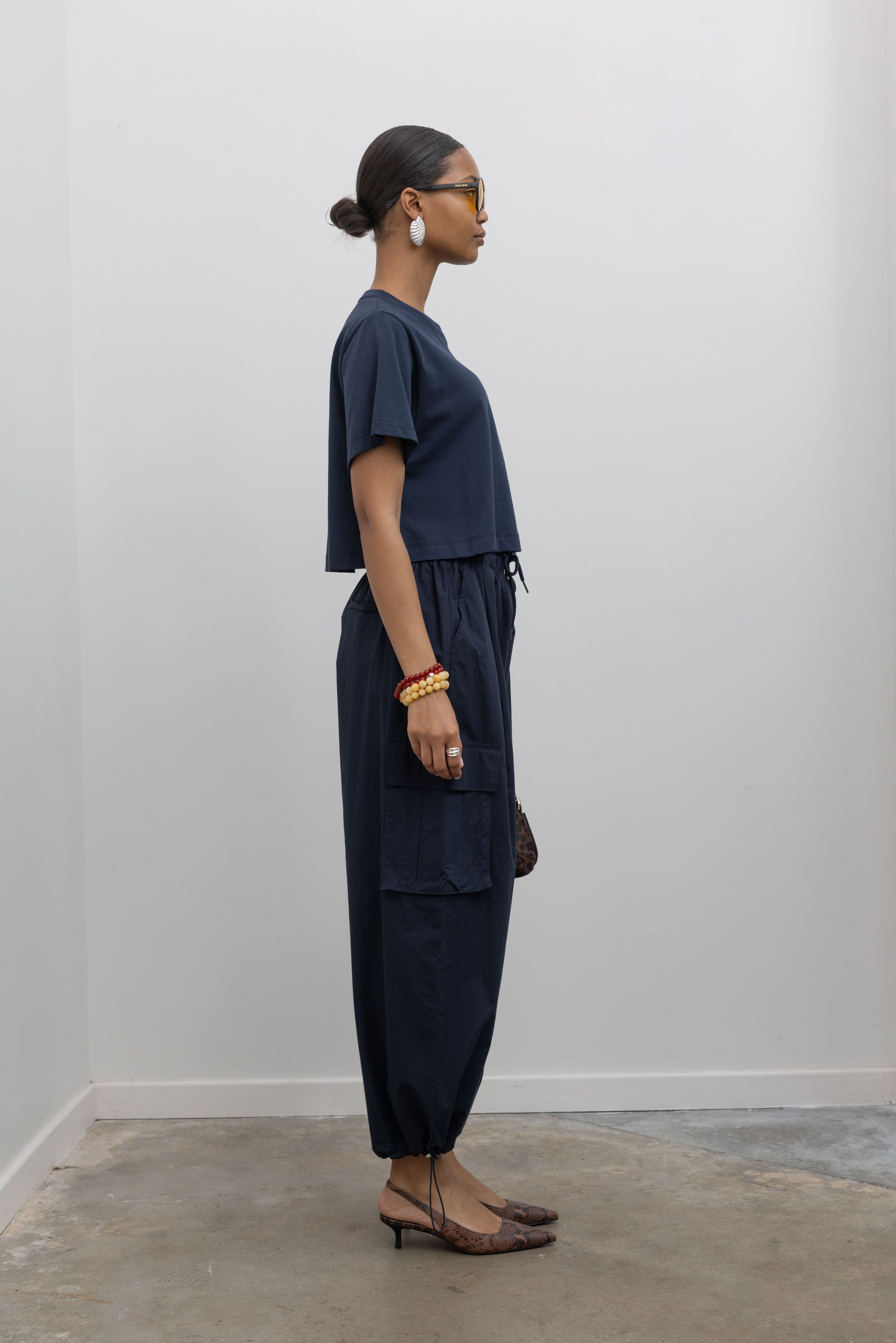 Side view of a Woman standing wearing capsule wardrobe essential in navy blue relaxed cropped t-shirt and a pair of 
navy lightweight cotton wide leg cargo pants. Featuring an oversized fit with drawstring ankles, a tie waist, and extra pockets on the legs at Alas Eius