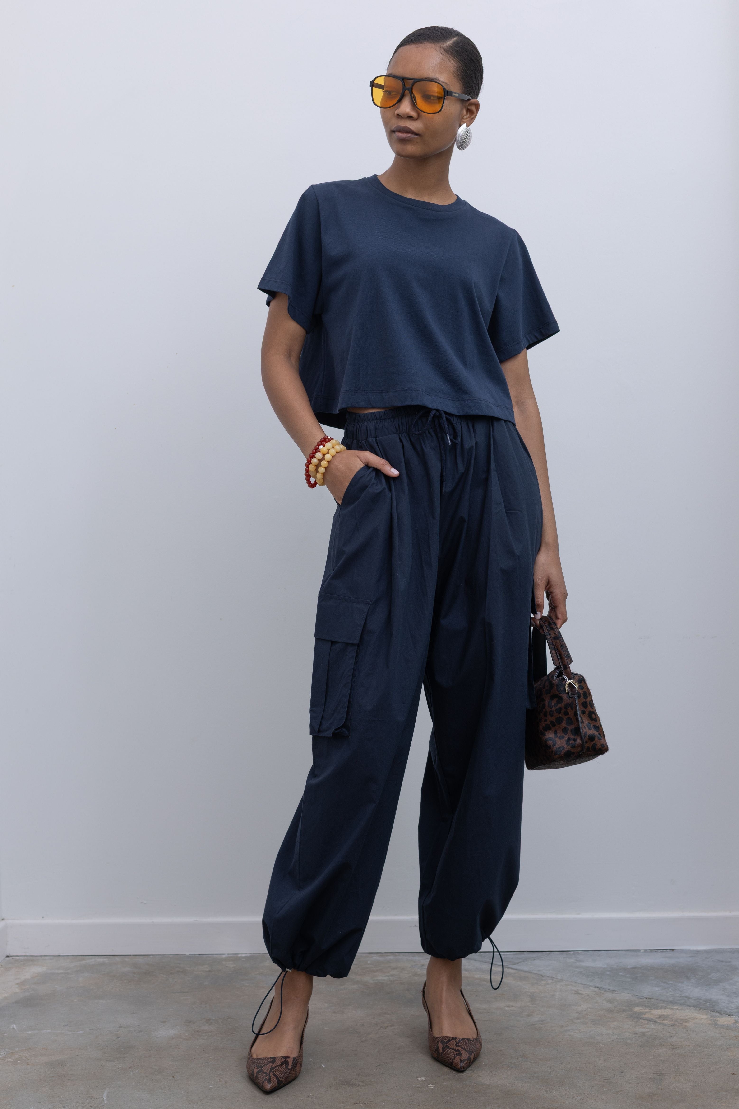 Woman standing posing with hand in side pocket wearing capsule wardrobe essential outfit in navy blue relaxed cropped t-shirt and a pair of 
Alas Eius Label navy blue lightweight cotton wide leg cargo pants that feature an oversized fit with drawstring cord ankles, a tie waist, and extra pockets on the legs at Alas Eius.