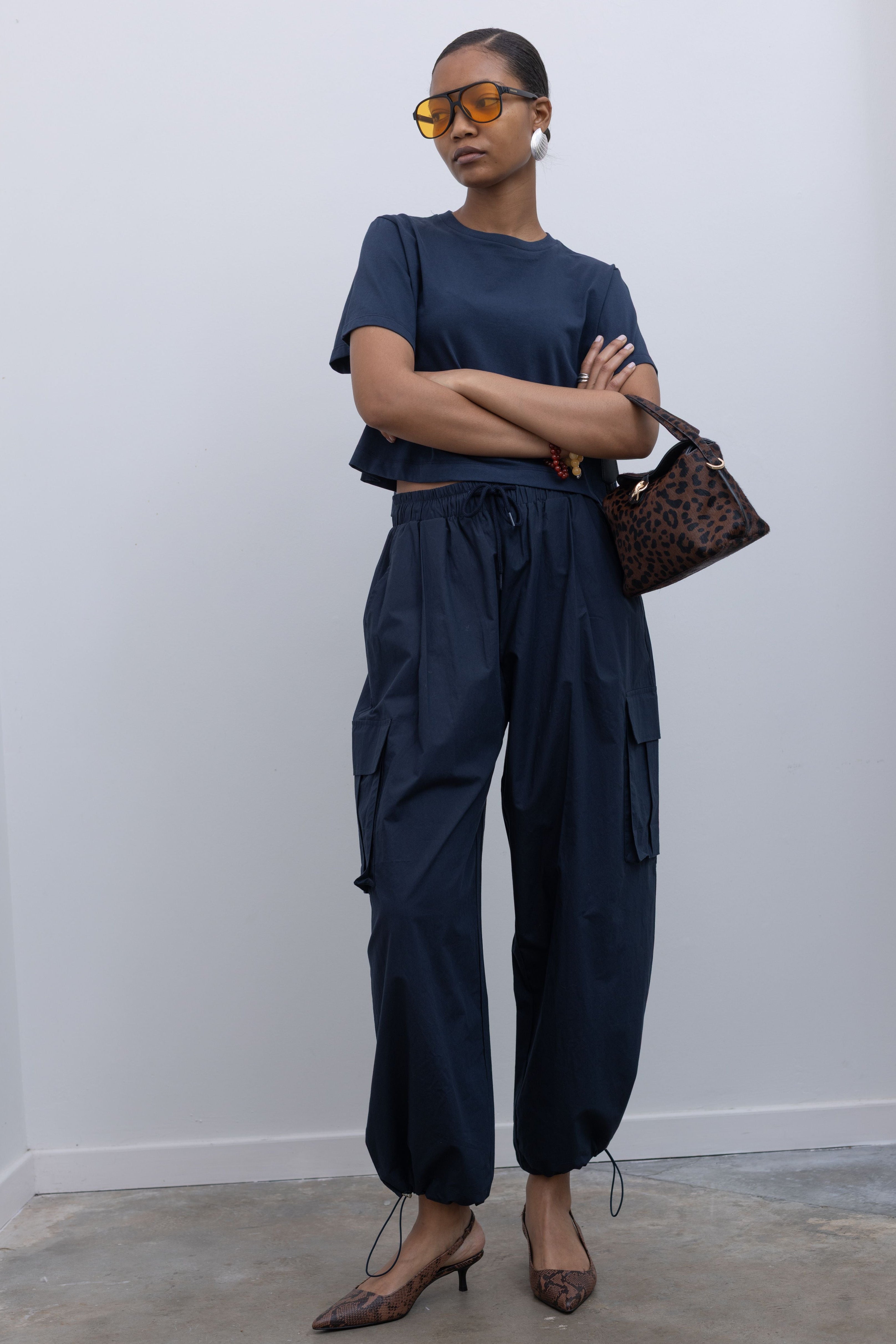 Woman standing with folded arms wearing capsule wardrobe essential in navy blue relaxed cropped short sleeved t-shirt and a pair of 
Alas Eius navy blue lightweight cotton wide leg cargo pants. Featuring an oversized fit with drawstring ankles, a tie waist, and extra pockets on the legs at Alas Eius.