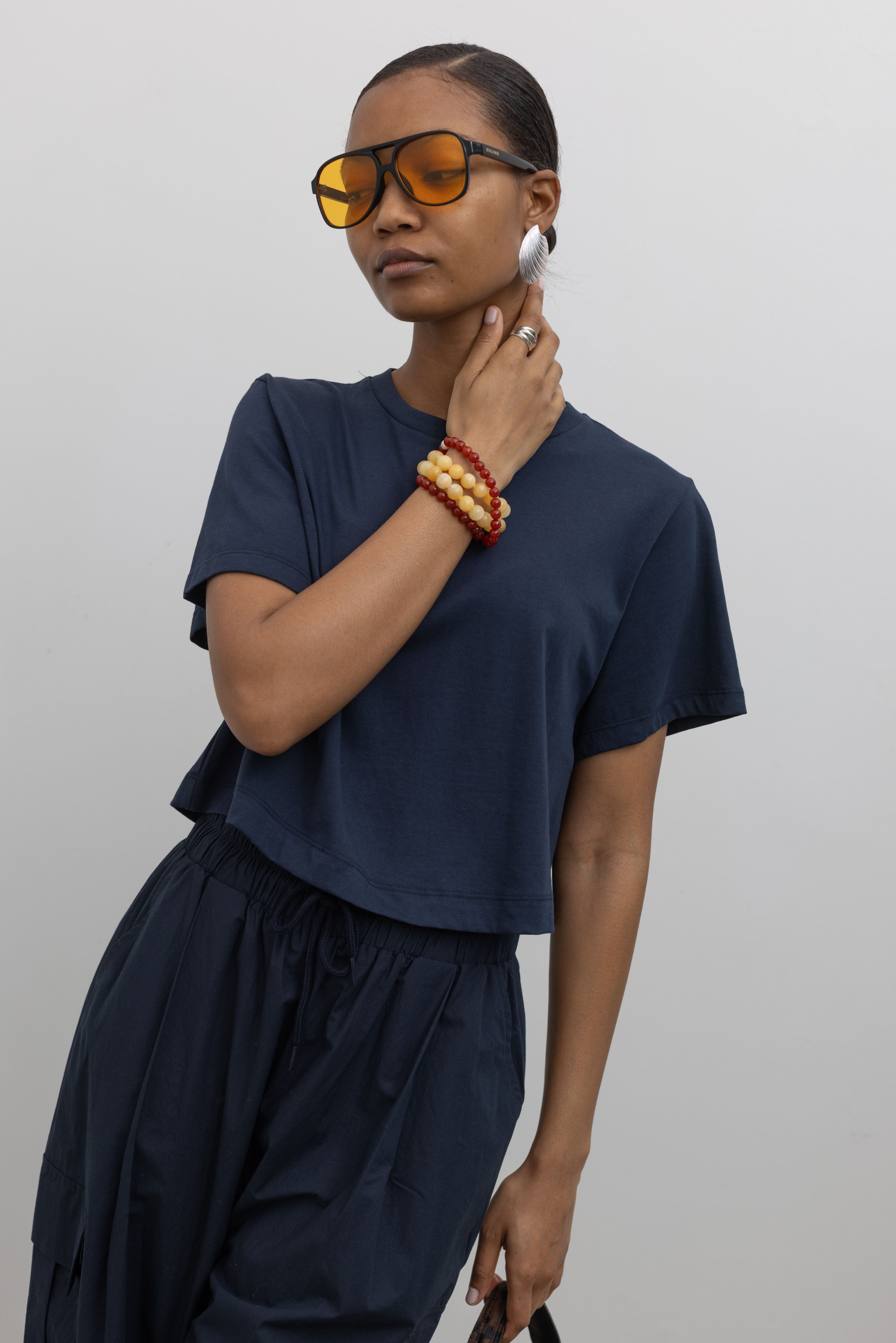 Detailed product view of woman standing wearing everyday essential Alas Eius Label Navy Blue Relaxed Cropped Short Sleeved T-Shirt with Navy Blue Cargo Pants and accessories at Alas Eius