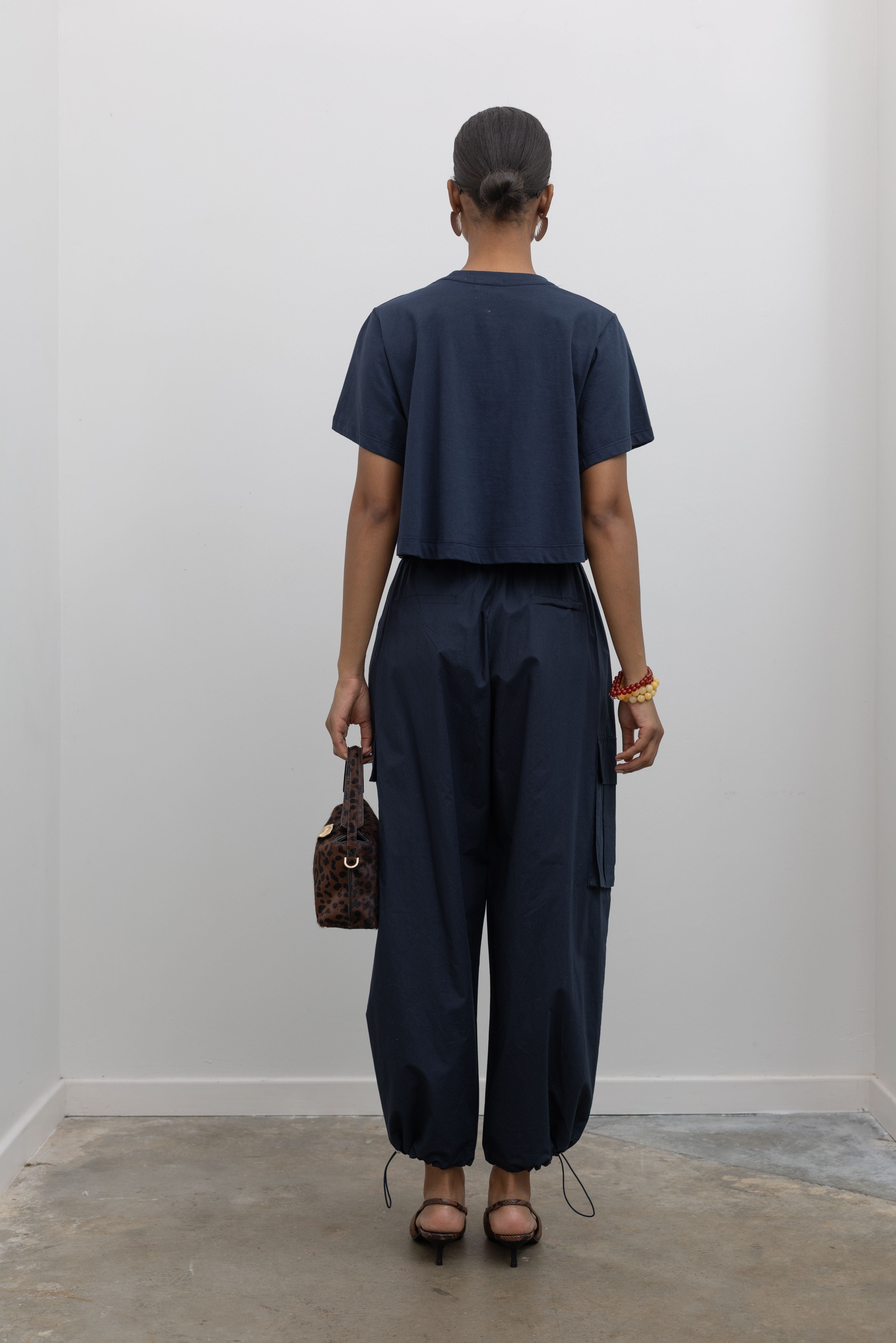 Back view of a Woman standing wearing capsule wardrobe essential in navy blue relaxed cropped t-shirt and a pair of navy blue lightweight cotton wide leg cargo pants. Featuring an oversized fit with drawstring ankles, a tie waist, and extra pockets on the legs at Alas Eius.