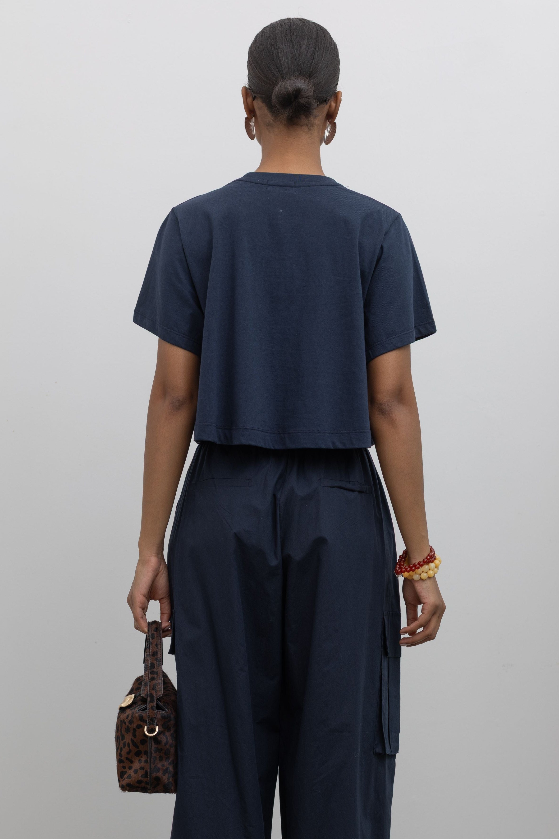 Detailed product back view of woman standing wearing everyday essential Alas Eius Label Navy Blue Relaxed Cropped Short Sleeved T-Shirt with Navy Blue Cargo Pants and accessories at Alas Eius