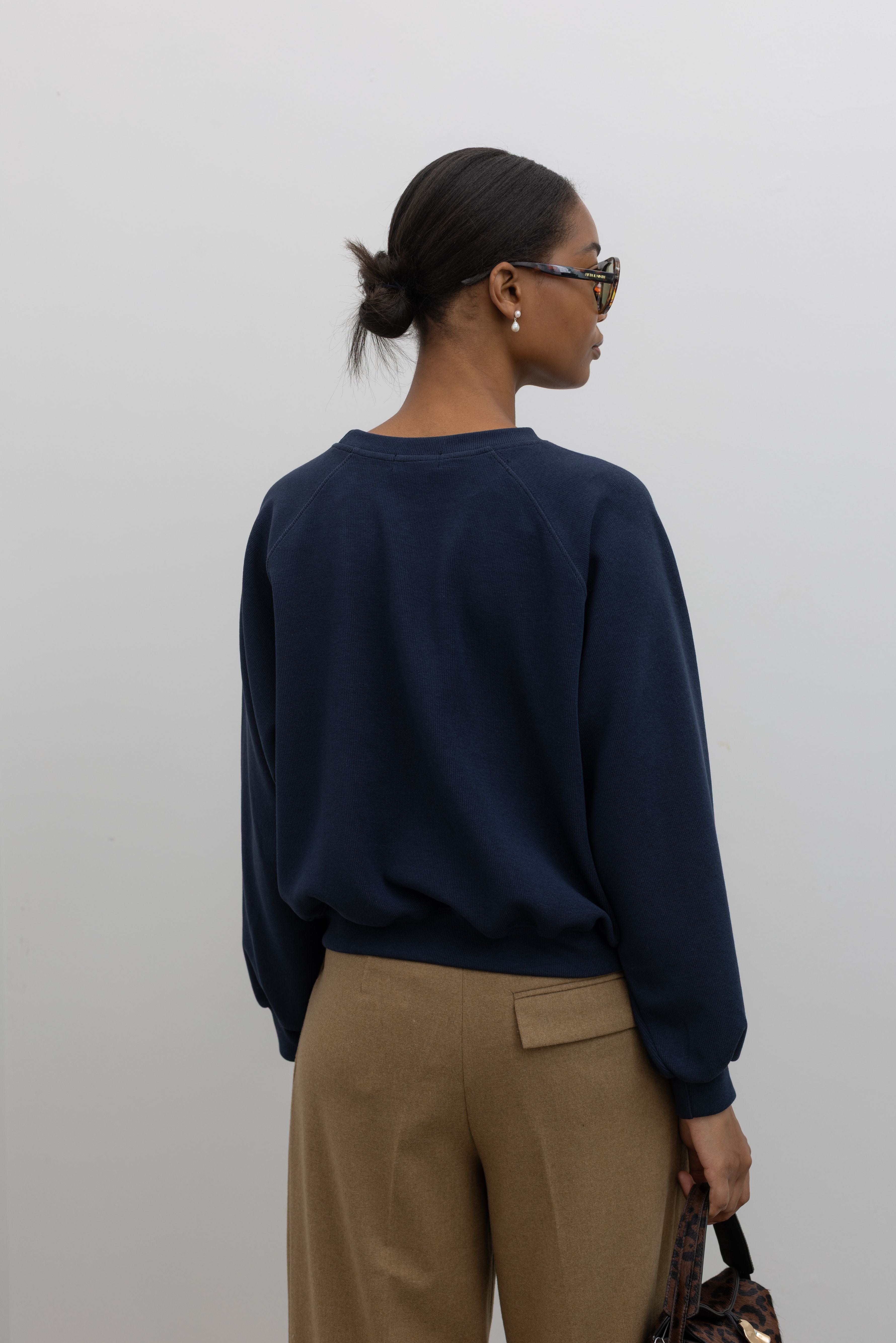Back view of a woman standing wearing a capsule wardrobe essential style in an Alas Eius label navy blue raglan sleeve elevated sweatshirt with a slightly cropped hem, landing at the waistband. 