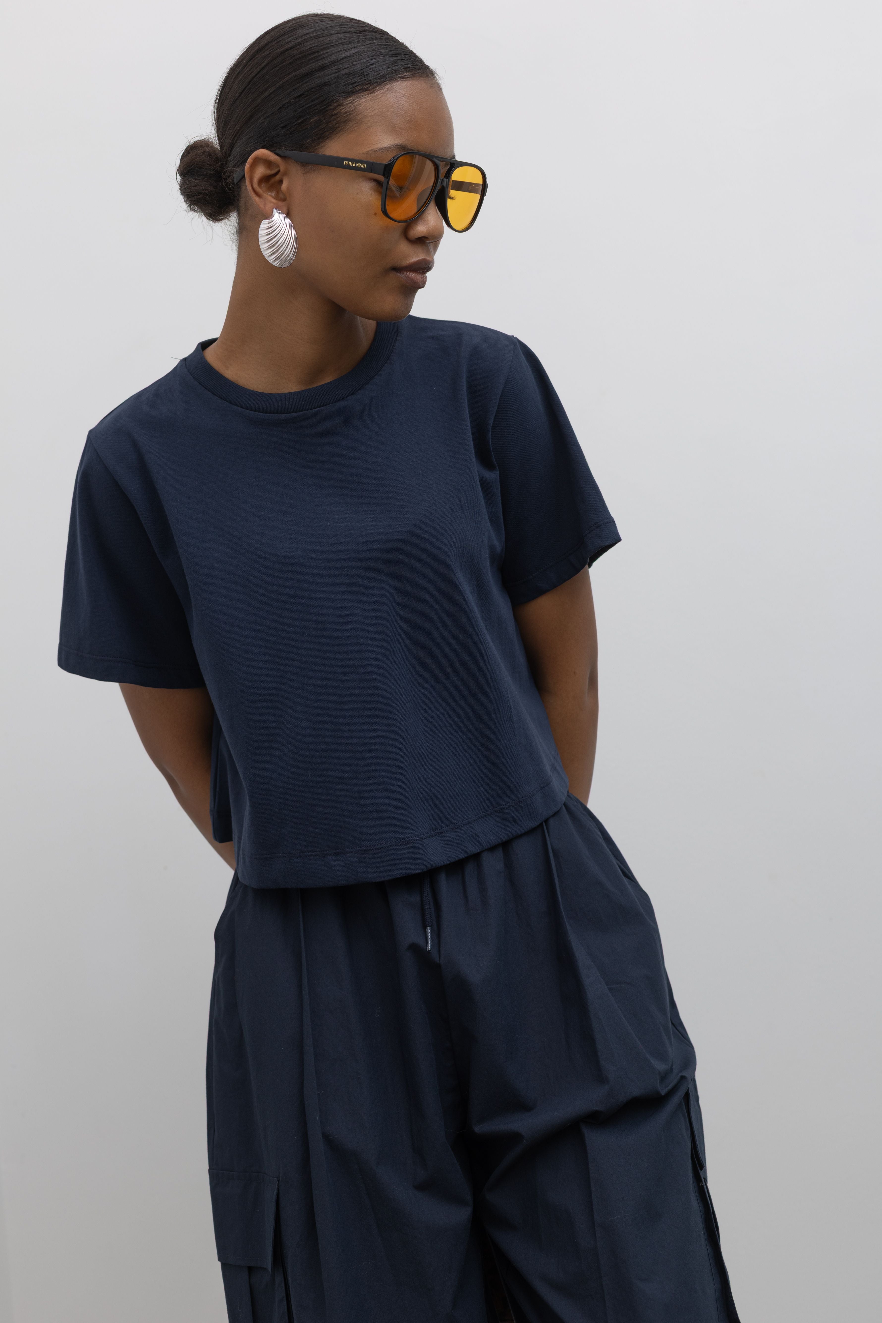 Detailed product front view of woman standing wearing everyday essential Alas Eius Label Navy Blue Relaxed Cropped Short Sleeved T-Shirt with Navy Blue Cargo Pants and accessories at Alas Eius