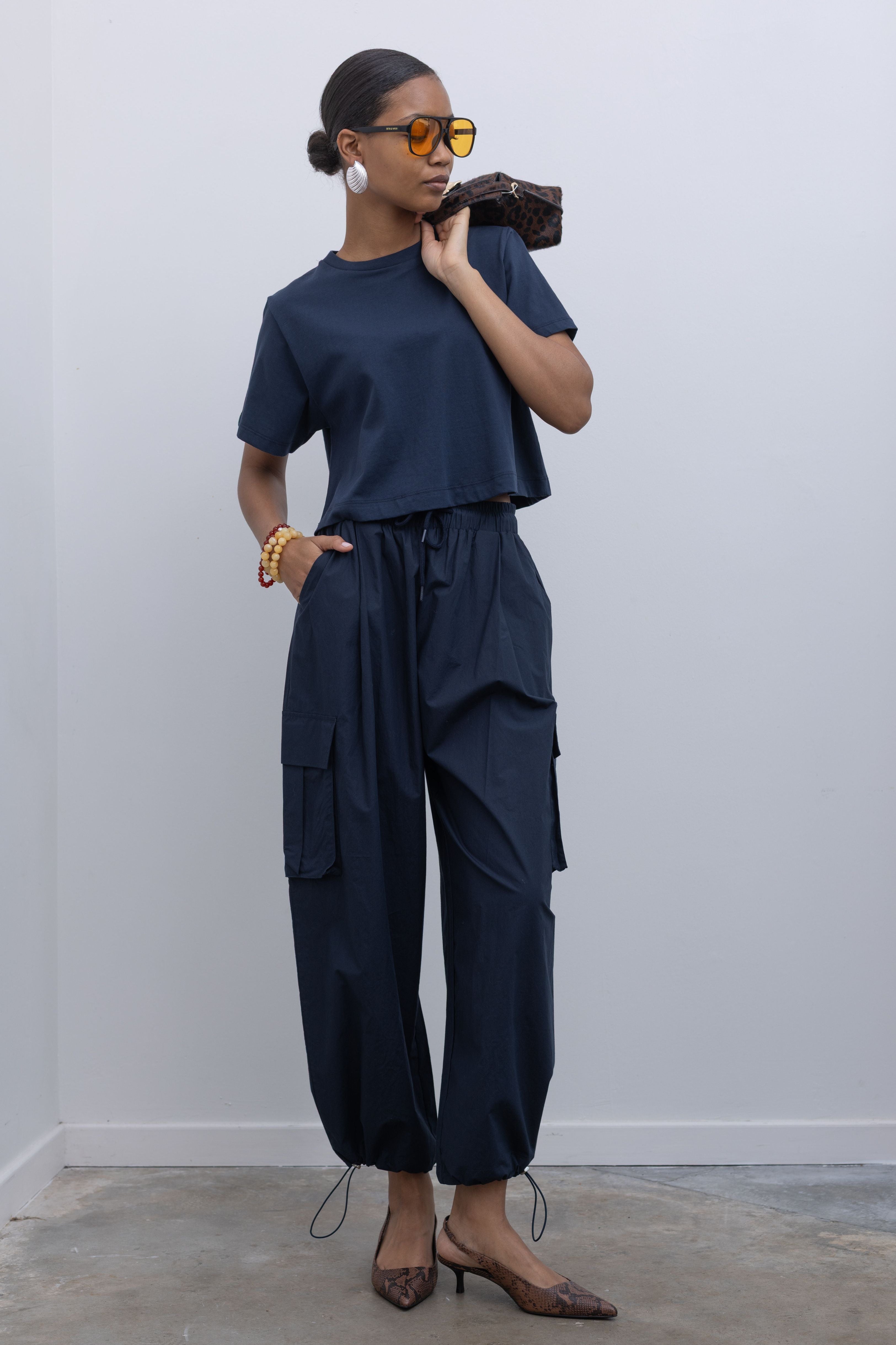 Woman standing wearing capsule wardrobe essential outfit in navy blue relaxed cropped t-shirt and a pair of 
Alas Eius Label navy blue lightweight cotton wide leg cargo pants. Featuring a relaxed fit with drawstring ankles, a tie waist, and extra pockets on the legs.