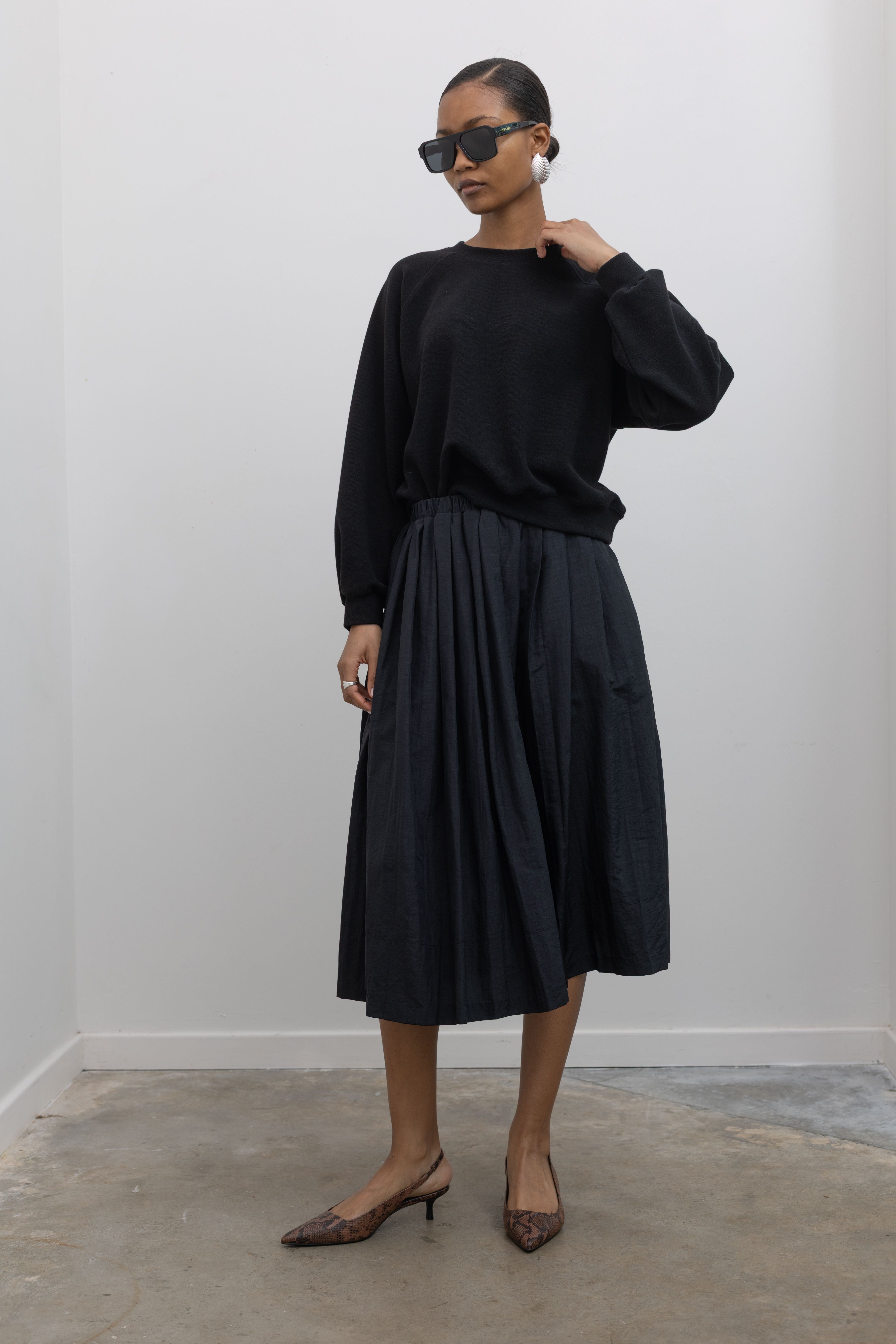 Front view of a woman posing wearing a capsule wardrobe outfit including a sophisticated Alas Eius Label black lightweight nylon pleated midi skirt, featuring a fully lined silhouette and finished with an elastic waistband and convenient side pockets. 