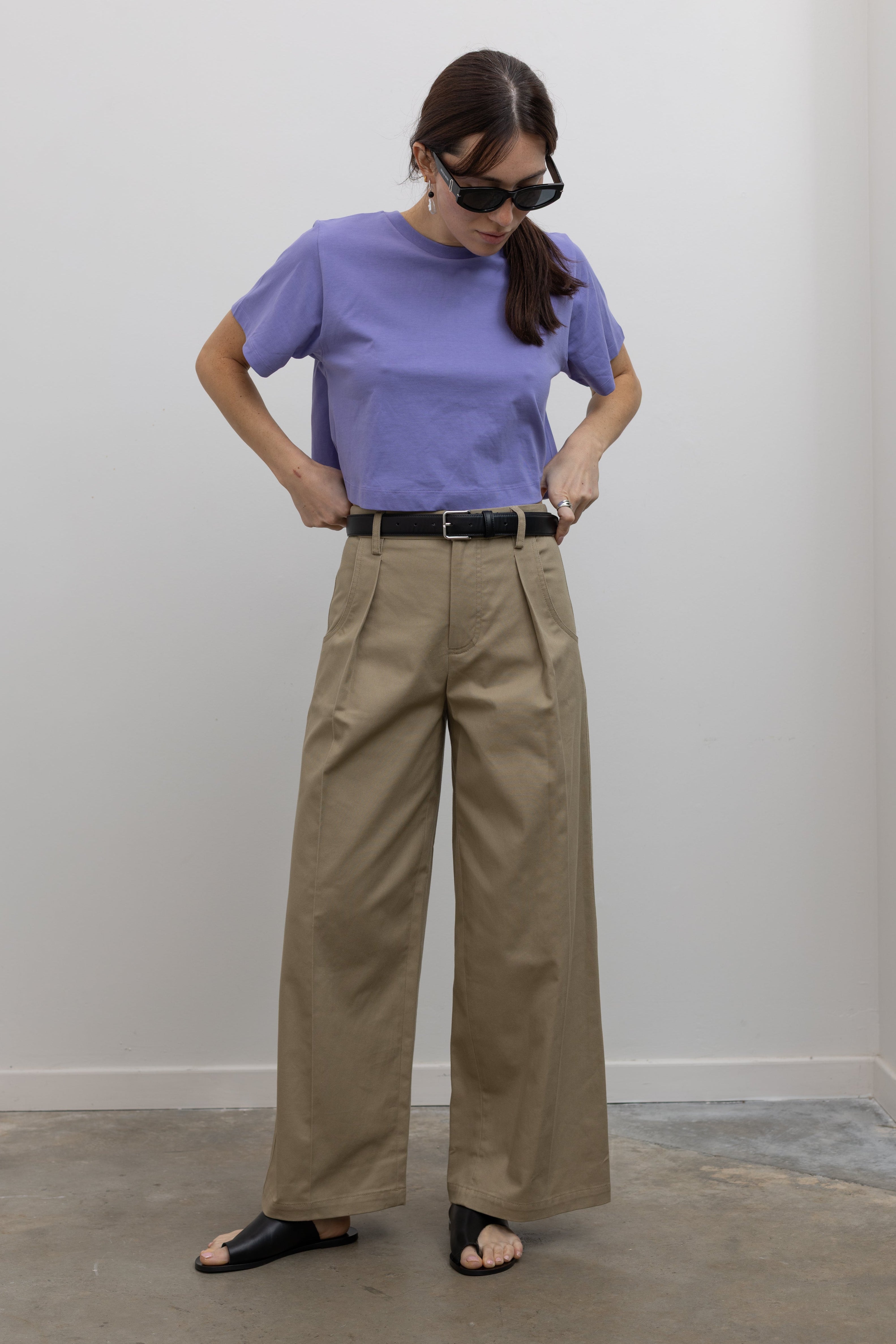 Woman standing wearing must-have capsule wardrobe essential in an Alas Eius Label Lavender Relaxed Boxy Cropped T-Shirt with khaki pleated high waisted trousers and thin black leather belt with silver hardware and black leather cutout sandals at Alas Eius 